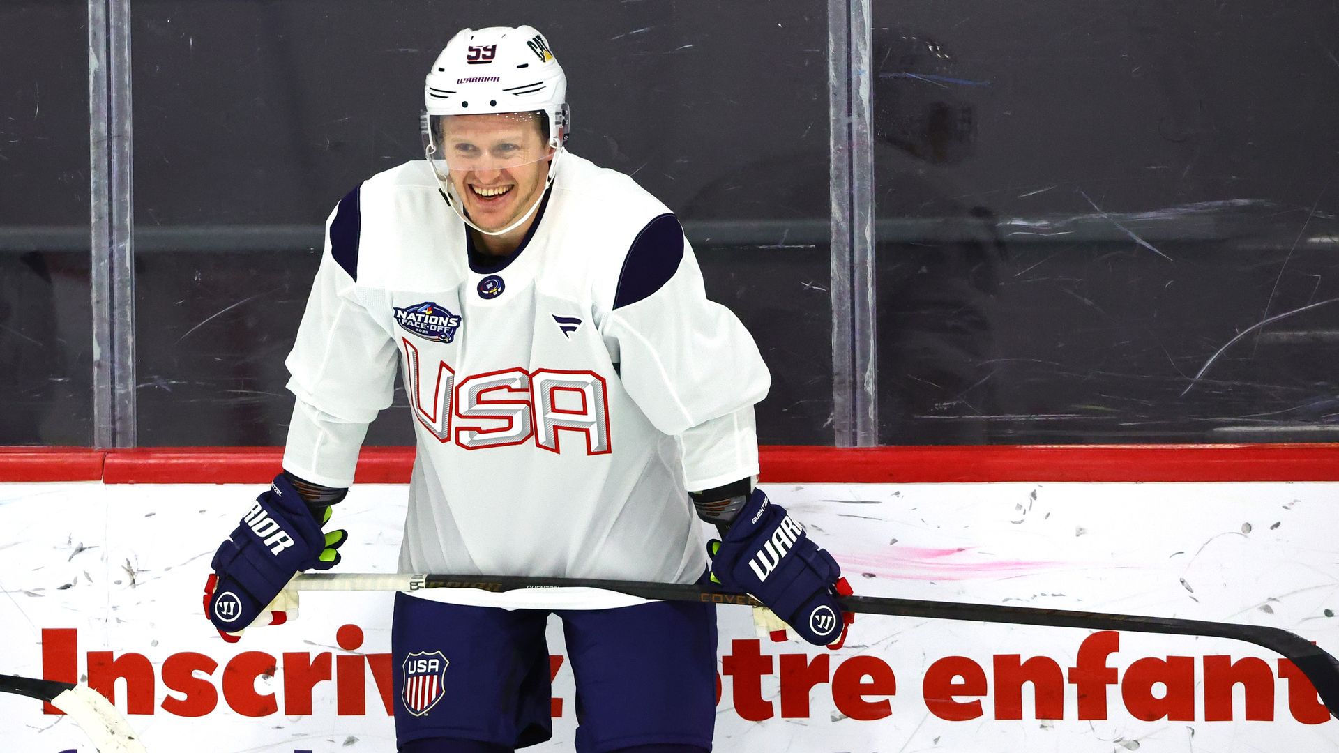 A hockey player in a white Team USA jersey stands and holds his stick across his waist during a practice