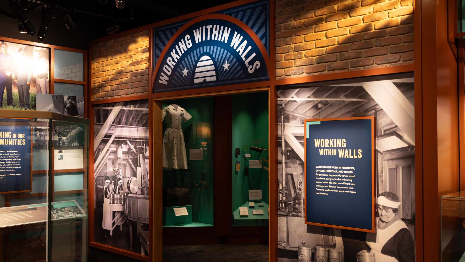Museum exhibit with brick walls and orange frames, a circular sign reading "WORKING WITHIN WALLS", blue panels, black-and-white factory photos, glass display cases, and a mannequin in a uniform.