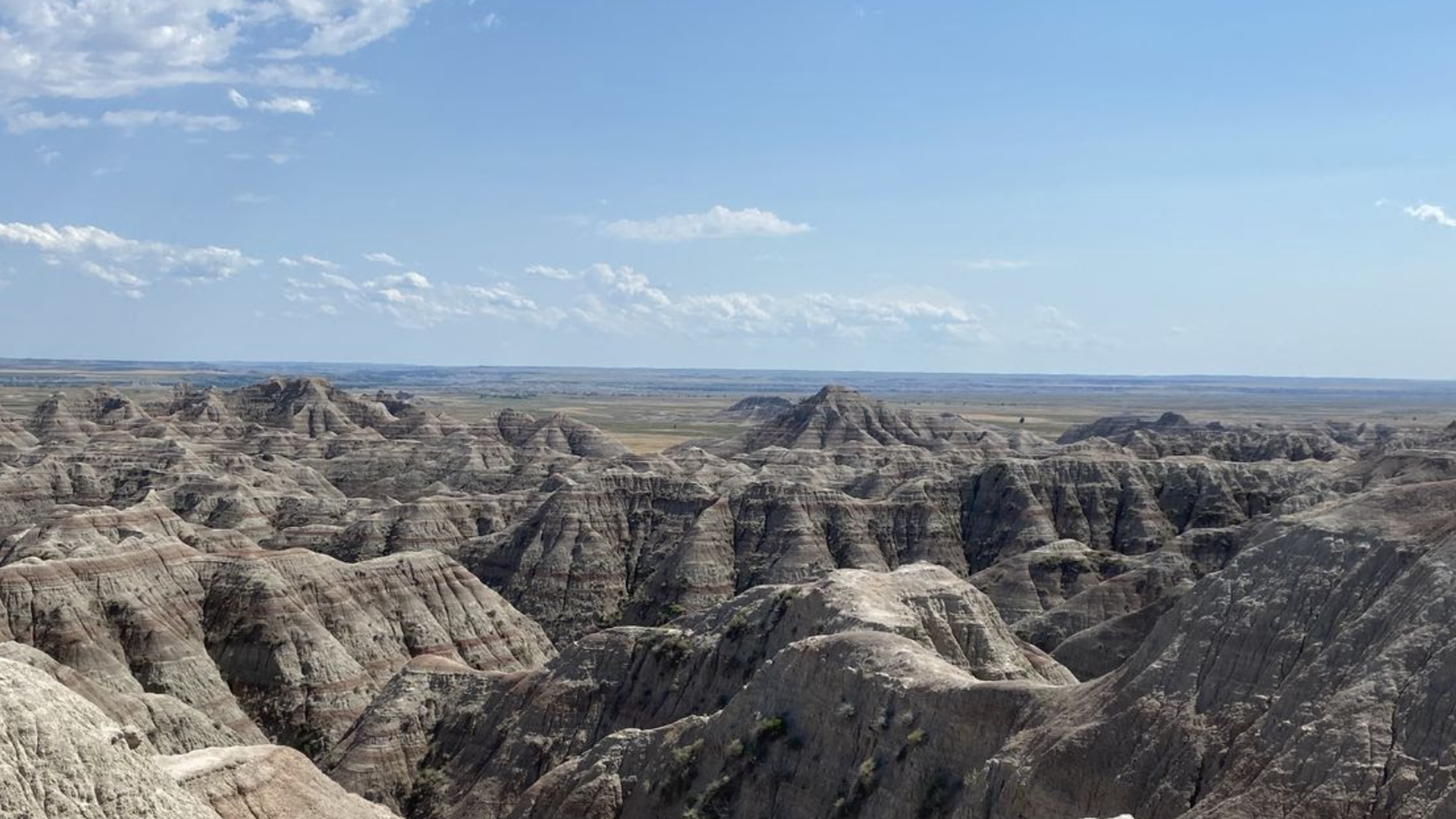 Expansive badlands with jagged, layered sedimentary formations in tan and gray, stretching to the horizon under a bright blue sky with scattered white clouds.