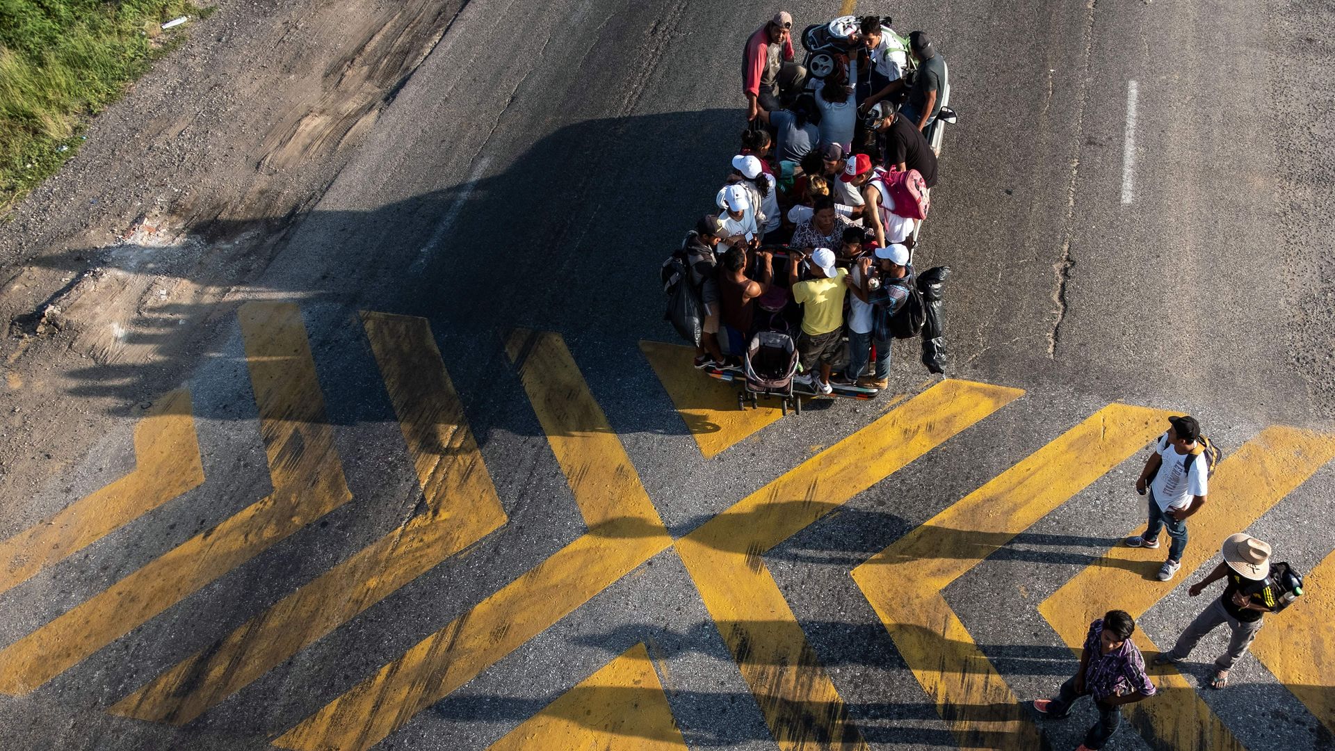An aerial shot of a truck on a road with Central American migrants who are members of the caravan sitting on top of it