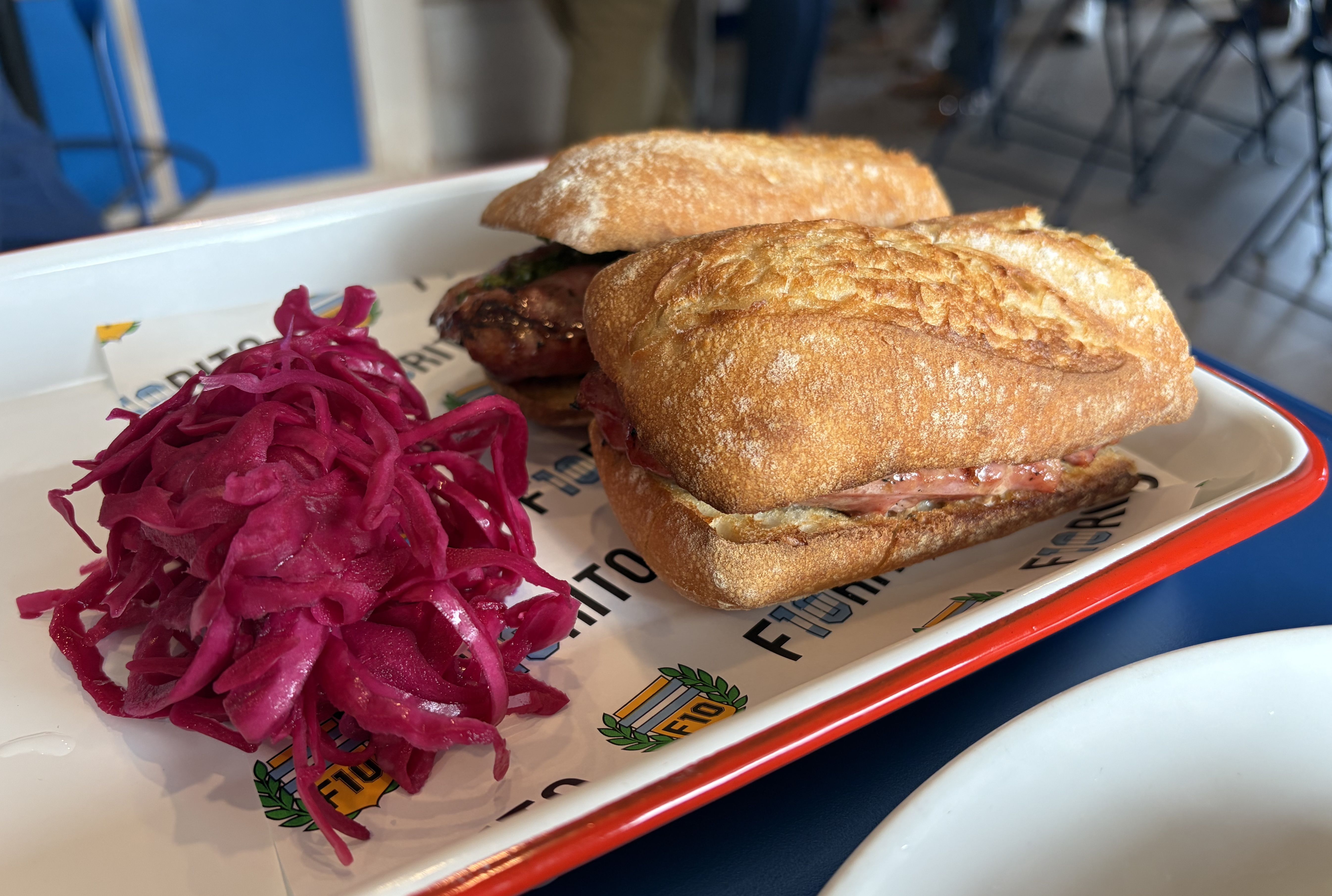 Two ciabatta sandwiches with meat on a white tray with red edges, served with a side of bright purple pickled cabbage in a casual dining setting.