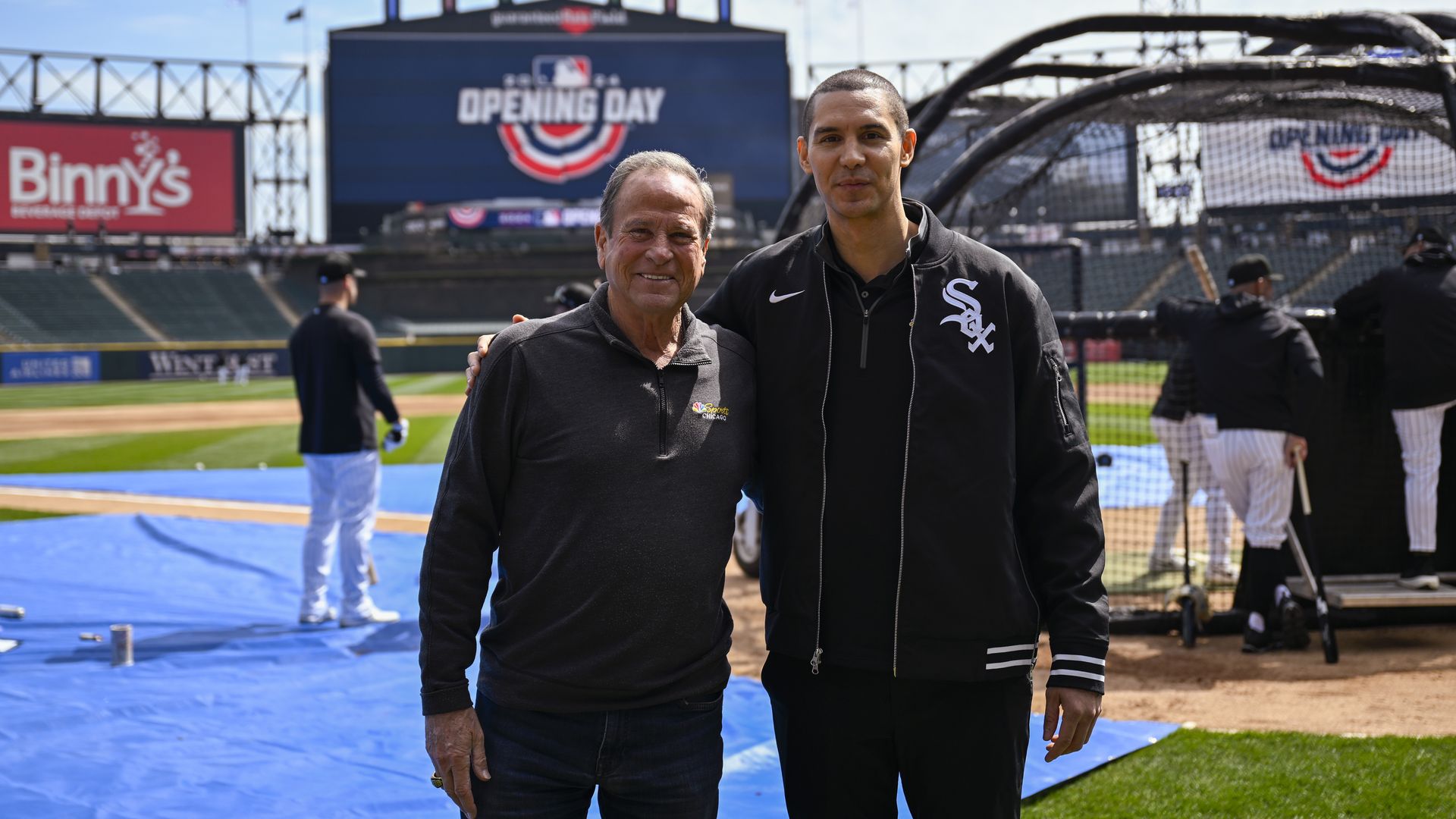 Two men stand on a baseball field, smiling. Behind them a large screen reads "Opening Day" with a red, white and blue emblem; a Binny's sign and a batting cage are visible.
