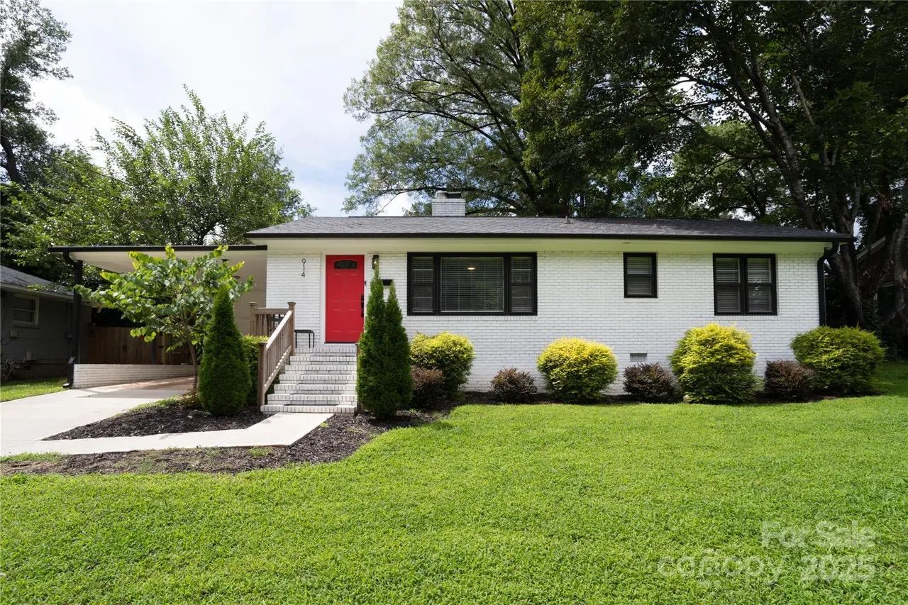 Single-story white brick house with black-trimmed windows, a bright red front door, surrounded by green bushes and grass, plus a concrete driveway and steps leading to the entrance.