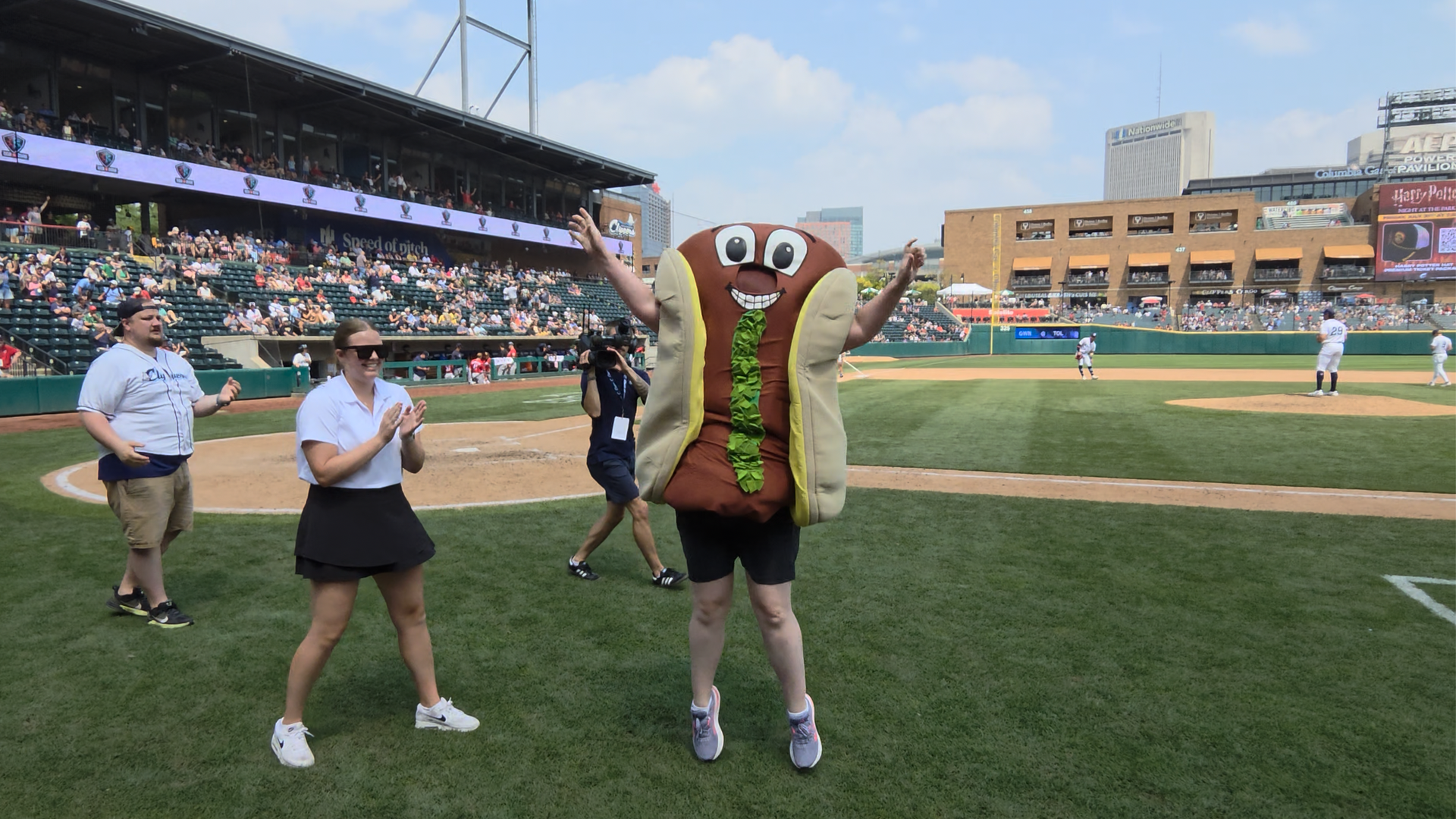 Alissa, in a hot dog costume with relish on it, raises her arms on a baseball field; two staffers clap nearby as a cameraman films, with a full stadium and city skyline in the background.