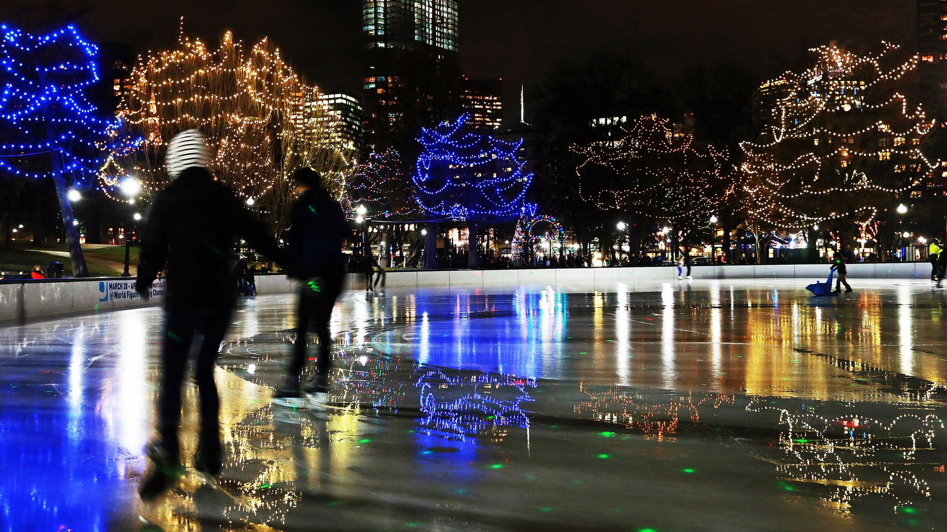 Two skaters in black clothes ice skating on a rink outside that's surrounded by lit up trees, white and blue lights, for the holidays.