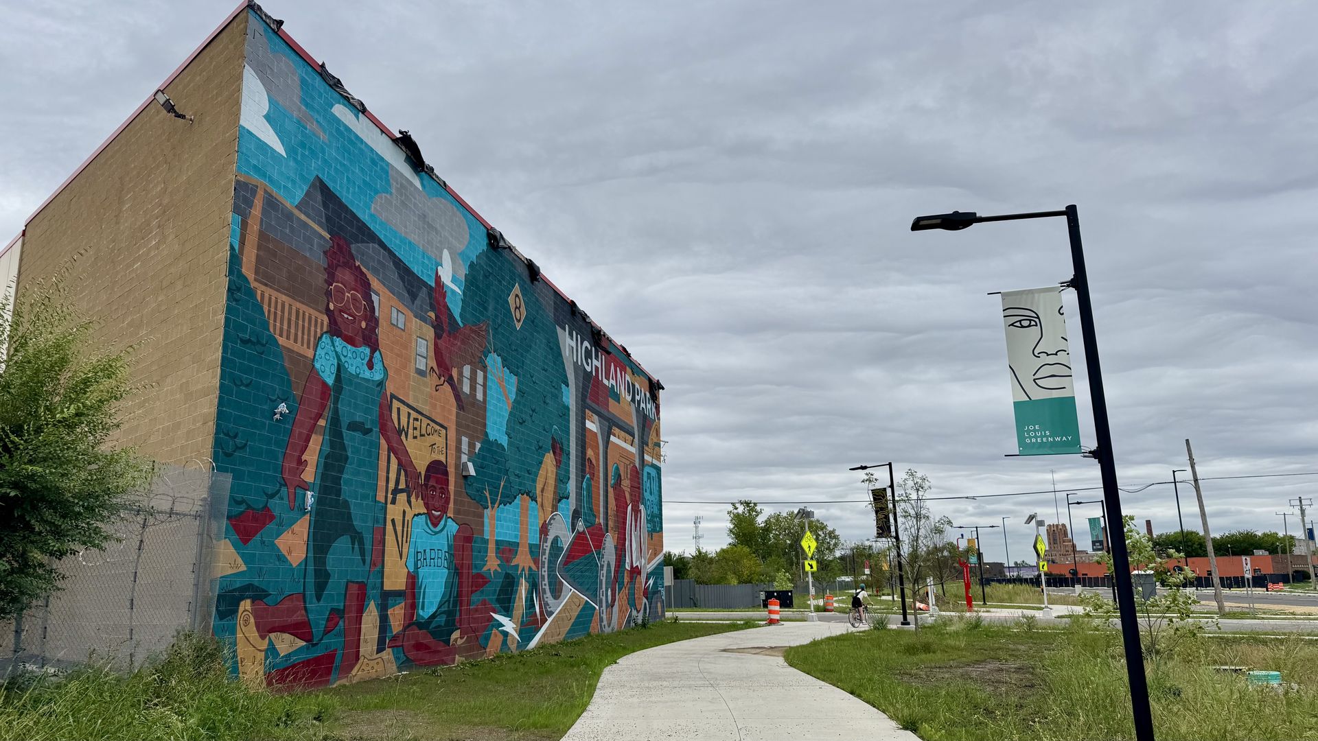A large mural on a brick building features colorful, stylized people and mountains in teal, brown, and red. A curved concrete path runs by a streetlamp with banners under a gray, cloudy sky.