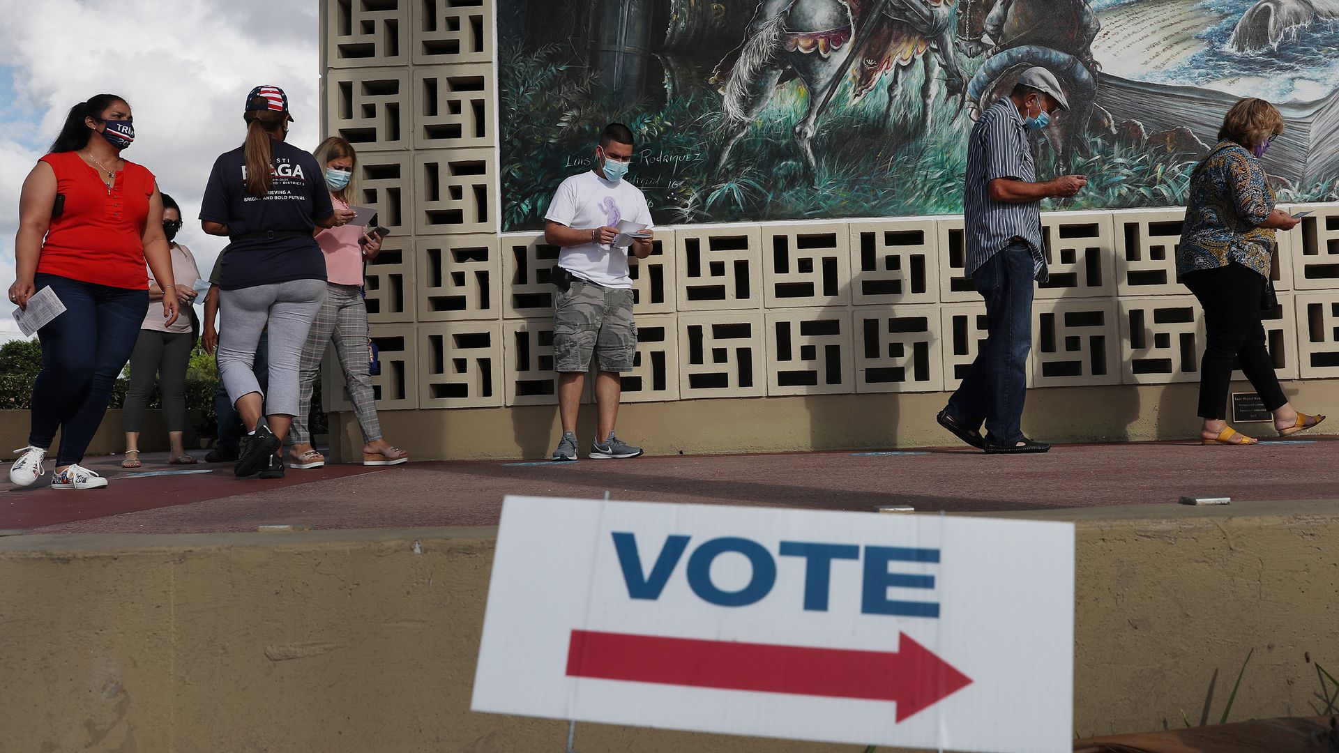 People waiting to vote outside spaced out and wearing masks. A blurred vote sign with an arrow is in the foregroud.