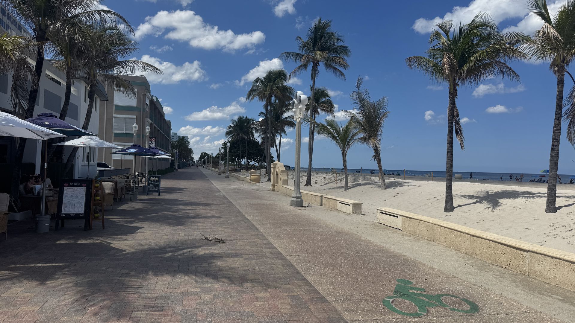 Sunny beachfront promenade with palm trees, brick walkway, and outdoor cafes on the left; beige buildings; sandy beach and blue sea on the right under a clear sky.