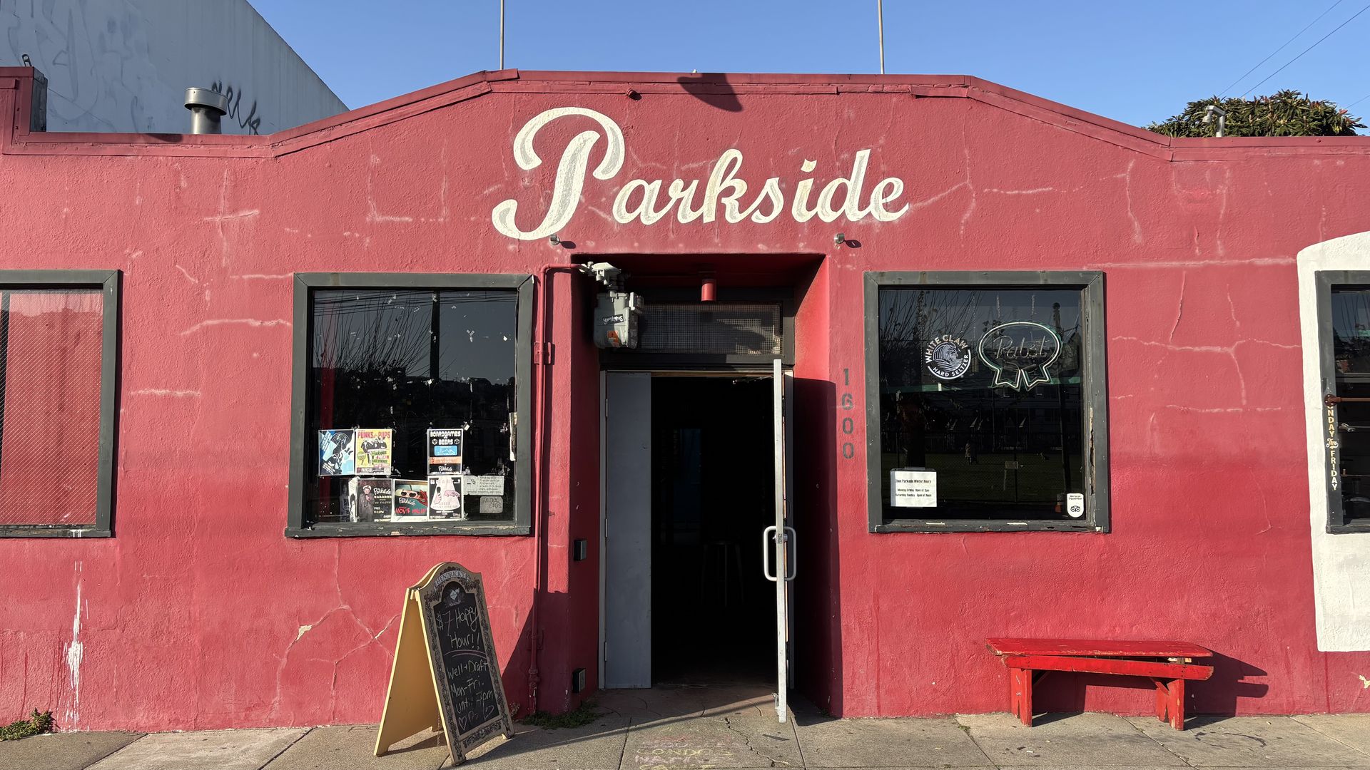 Front of a red building with "Parkside" painted above a partially open door, two large windows with posters, a chalkboard sign on the sidewalk, and a red bench under the right window.