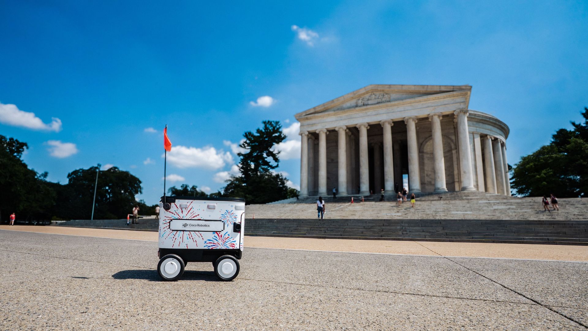 White Coco Robotics wheeled robot with colorful burst graphics sits on a paved plaza in front of a large neoclassical temple with tall columns under a bright blue sky.