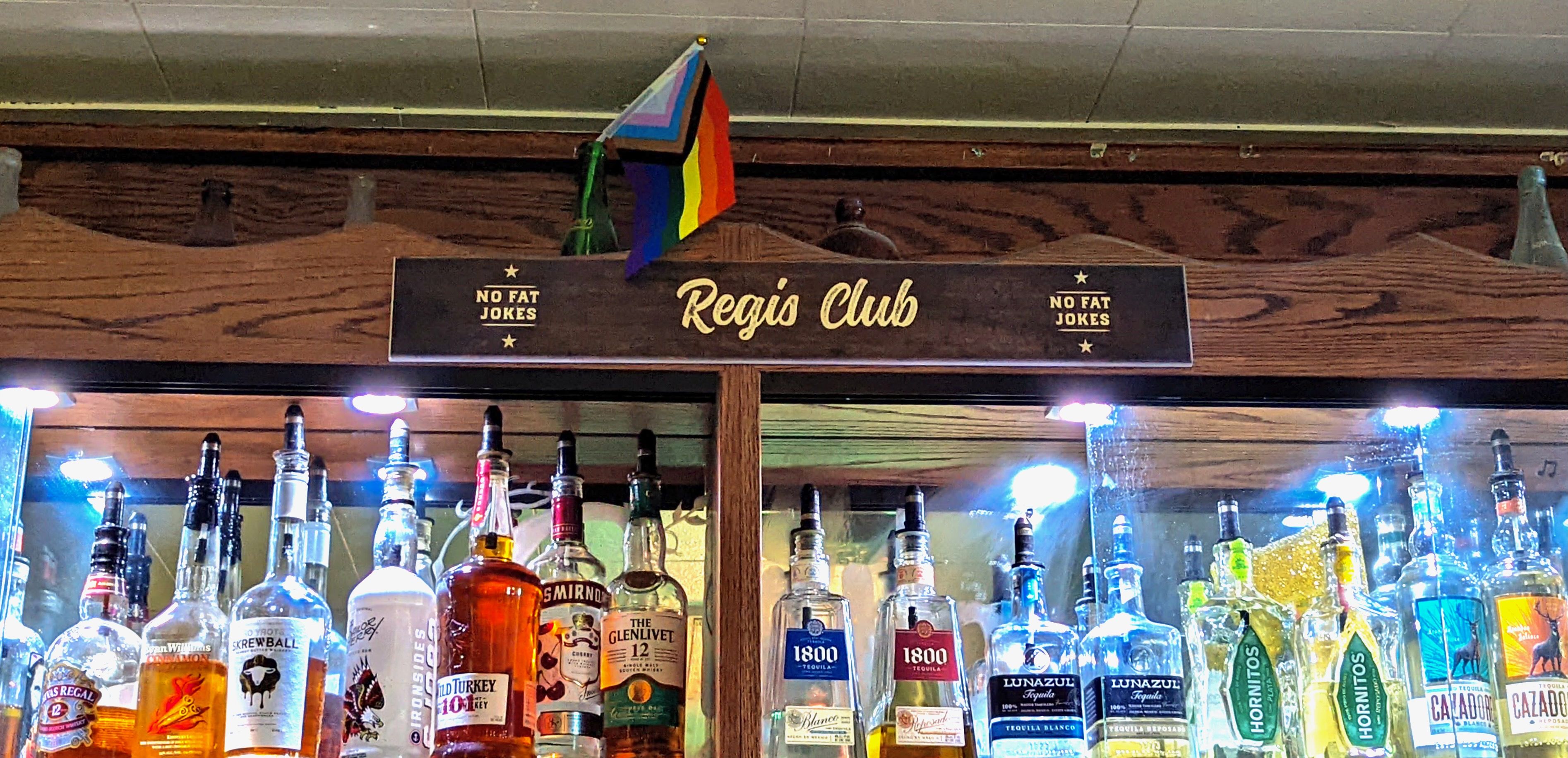 A sign reads "Regis Club: No fat jokes" with a pride flag hanging above it and bottles on a liquor cabinet below it. 