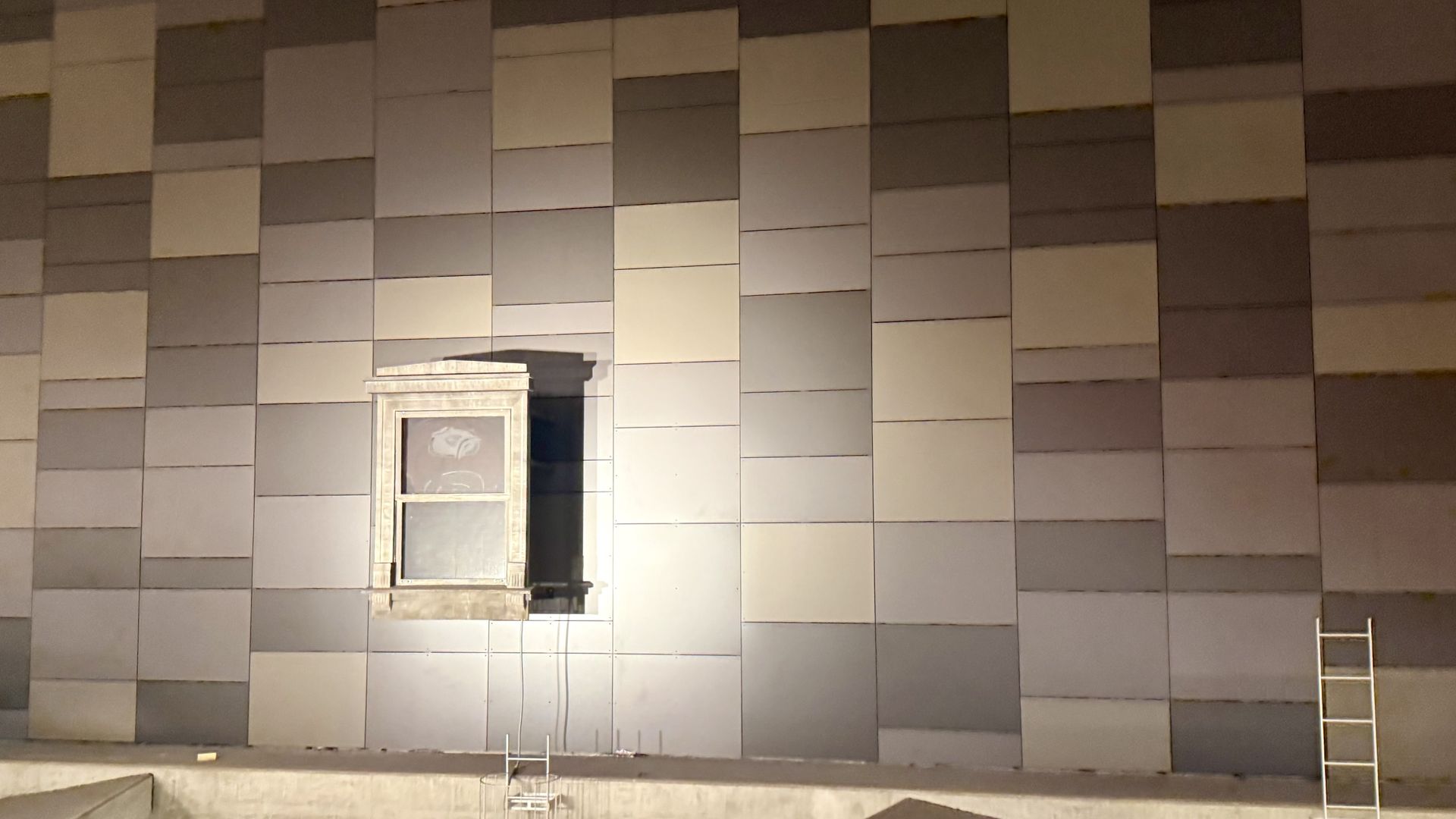 A tall wall covered with a patchwork of gray rectangular panels in various tones; a white-framed window sits near the lower left, with two metal ladders leaning against the wall.