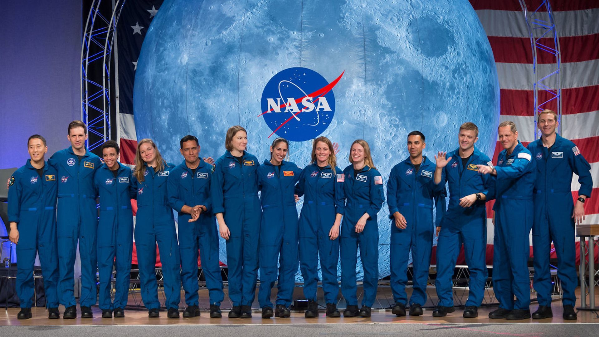 In this image, a line of NASA astronauts stand in front of the NASA logo and the moon