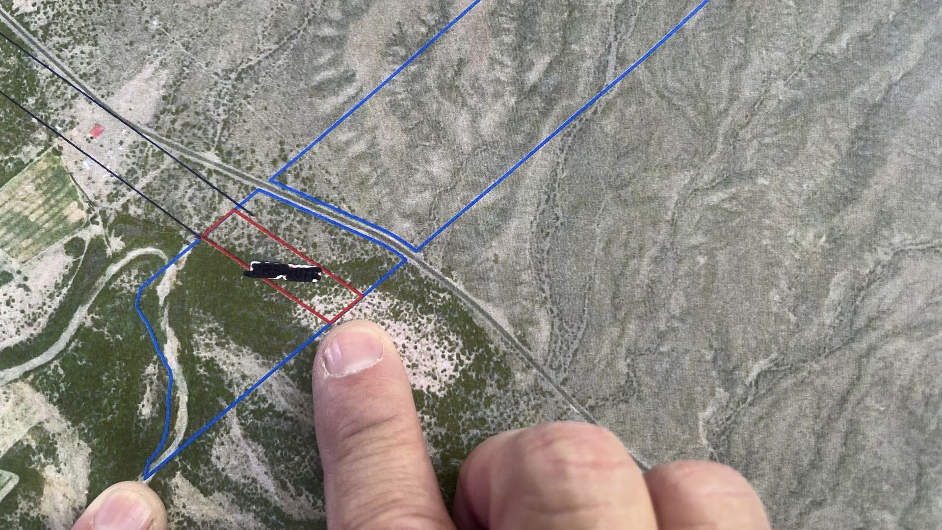 Top-down map shows blue property boundaries; a red rectangular lot inside a blue border is being pointed at by a finger. A small red structure, green patches left, and barren gray terrain right.