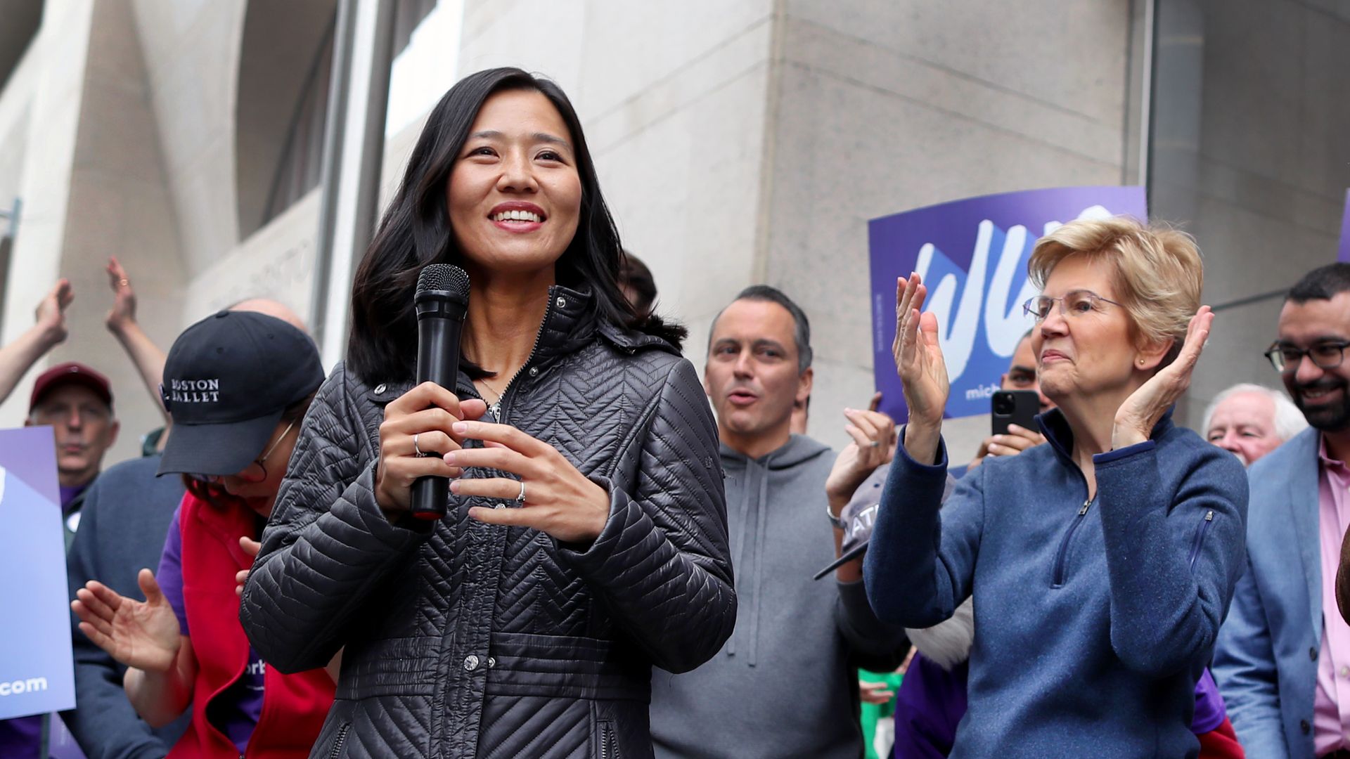 Photo of Michelle Wu smiling and holding a microphone as supporters including Elizabeth Warren clap around her