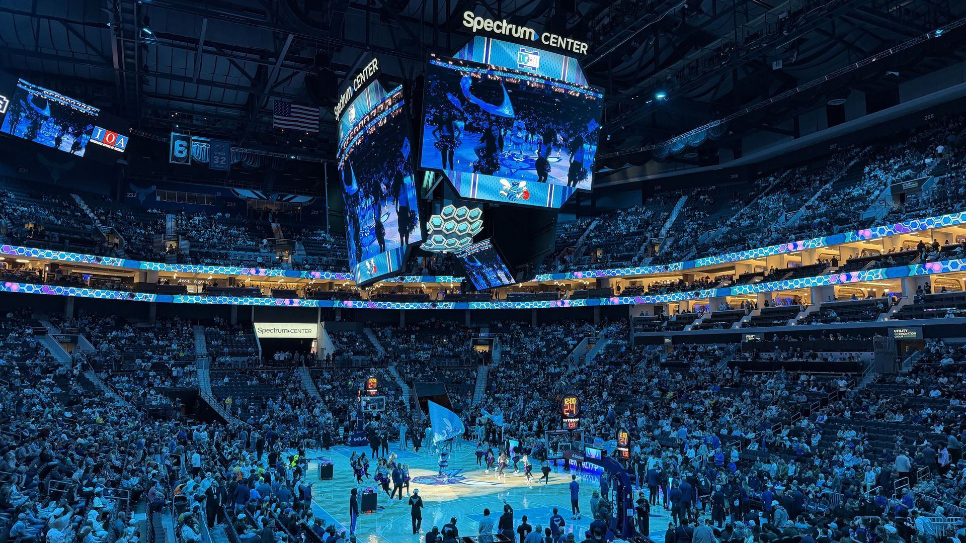 Inside Spectrum Center, a full arena of fans watches a basketball warm-up. A giant hanging scoreboard dominates the center as blue hexagon LED ribbons encircle the stands.