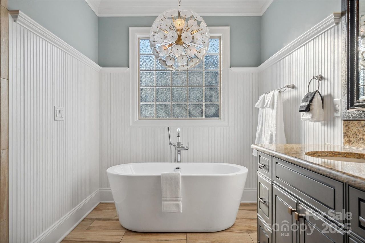 Modern bathroom with white freestanding tub, wood-look tile floor, pale blue walls with white wainscoting, decorative spherical chandelier, frosted window, and dark wood vanity with granite countertop.