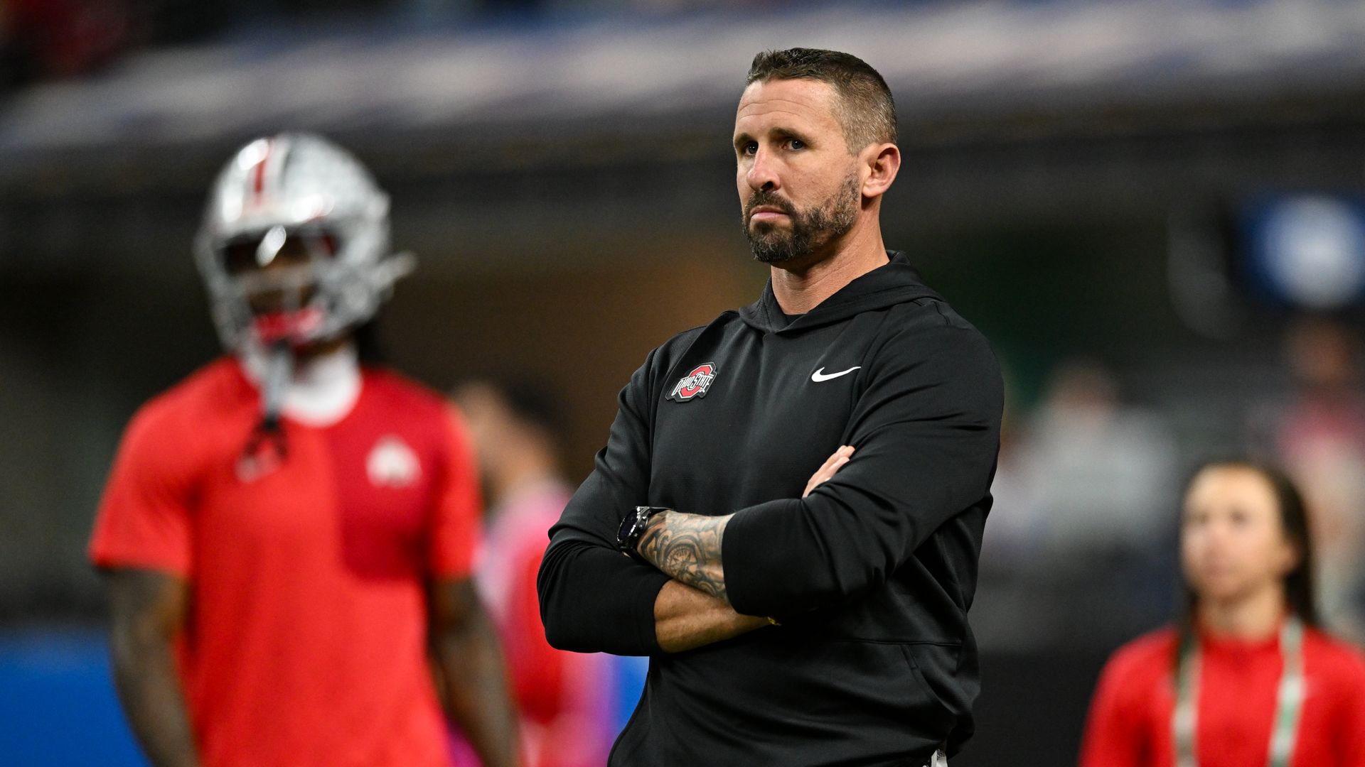 Ohio State offensive coordinator Brian Hartline stands with his arms crossed in a black hooded shirt prior to the Big Ten Championship football game last season.