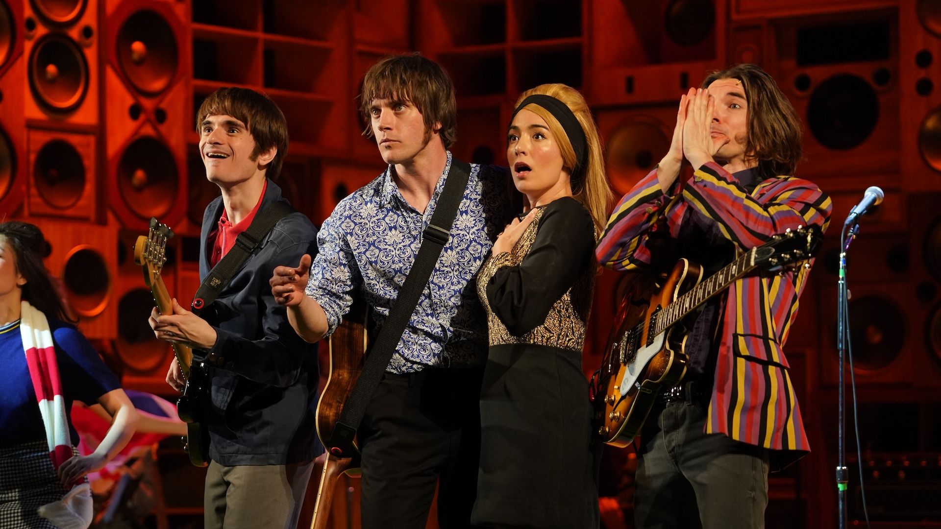 Four people in a row, two men holding guitars, a woman with a blond wig and a man with shoulder length hair holding a guitar.