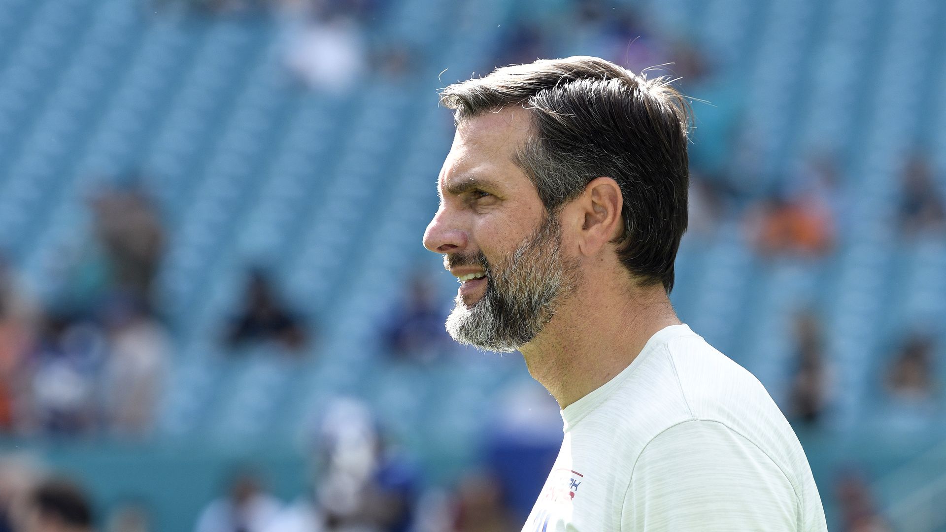 Side profile of Derek Dooley with short gray hair and beard, wearing a white Nike shirt, standing in a stadium with blurred blue seats and people in the background.