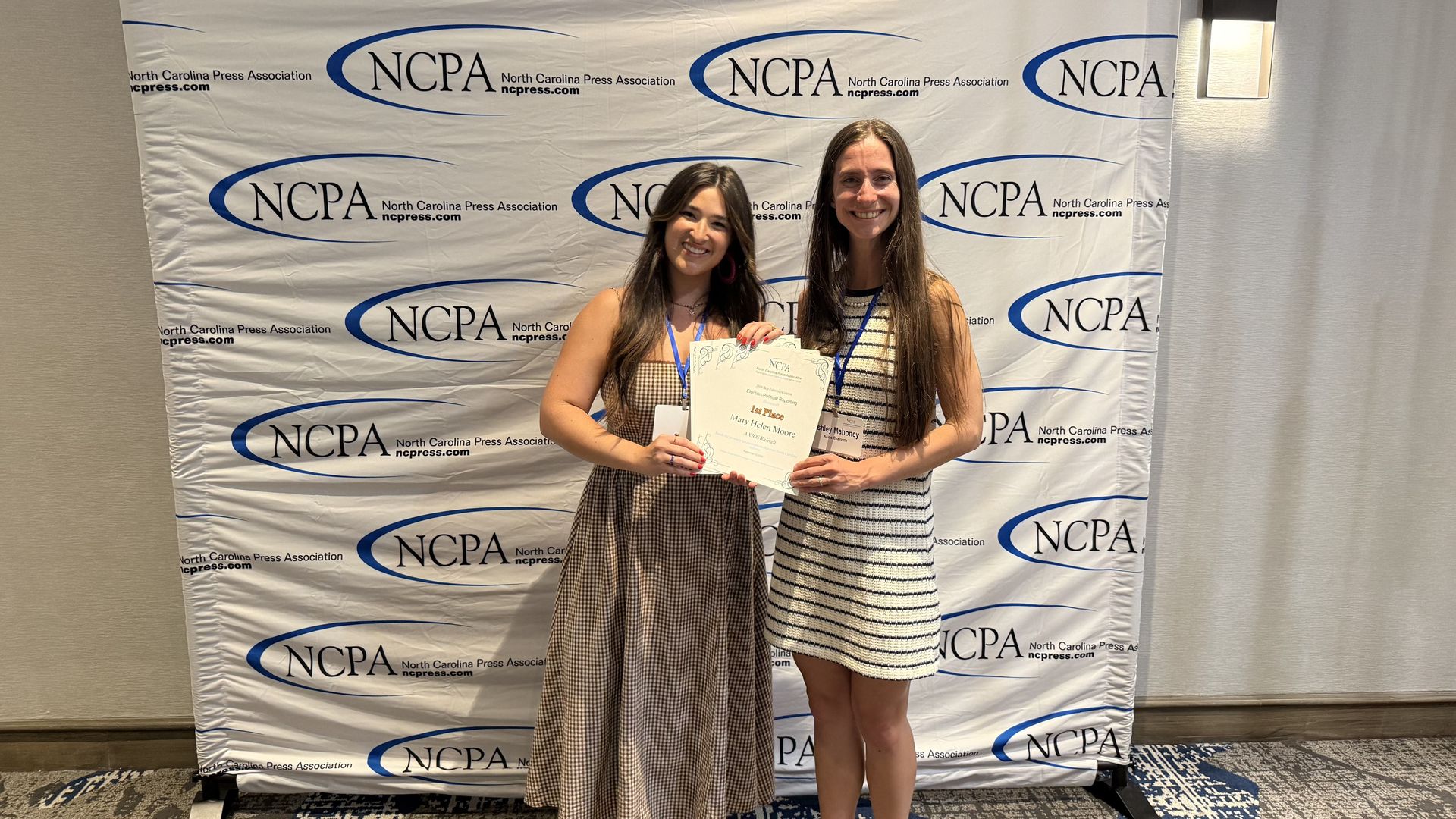Two smiling women stand in front of a white backdrop covered with blue NCPA logos, each holding a certificate. They wear light dresses; a wall sconce and patterned carpet are visible behind them.