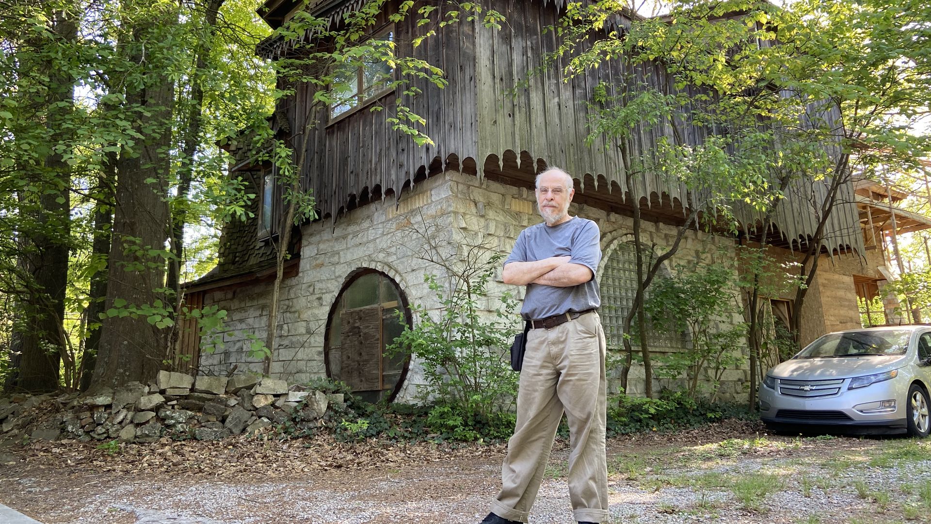 A man stands in front of a custom built house of stone and cedar siding with intricate woodwork