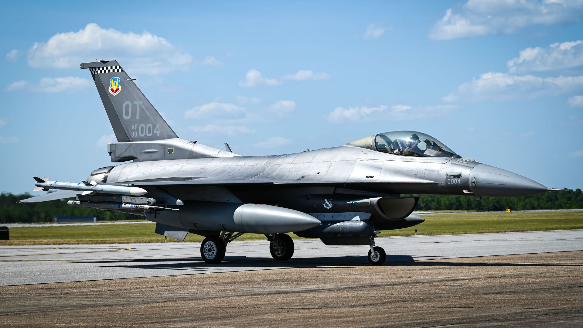 Side view of an F-16 on a runway. The pilot is visible in the cockpit. The sky overhead is blue.