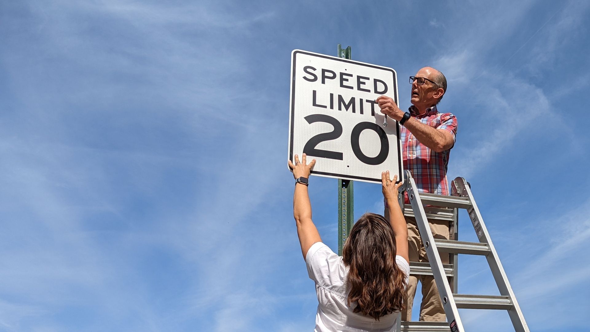 A man and a woman hang a street sign that advertises a speed limit of 20 mph.