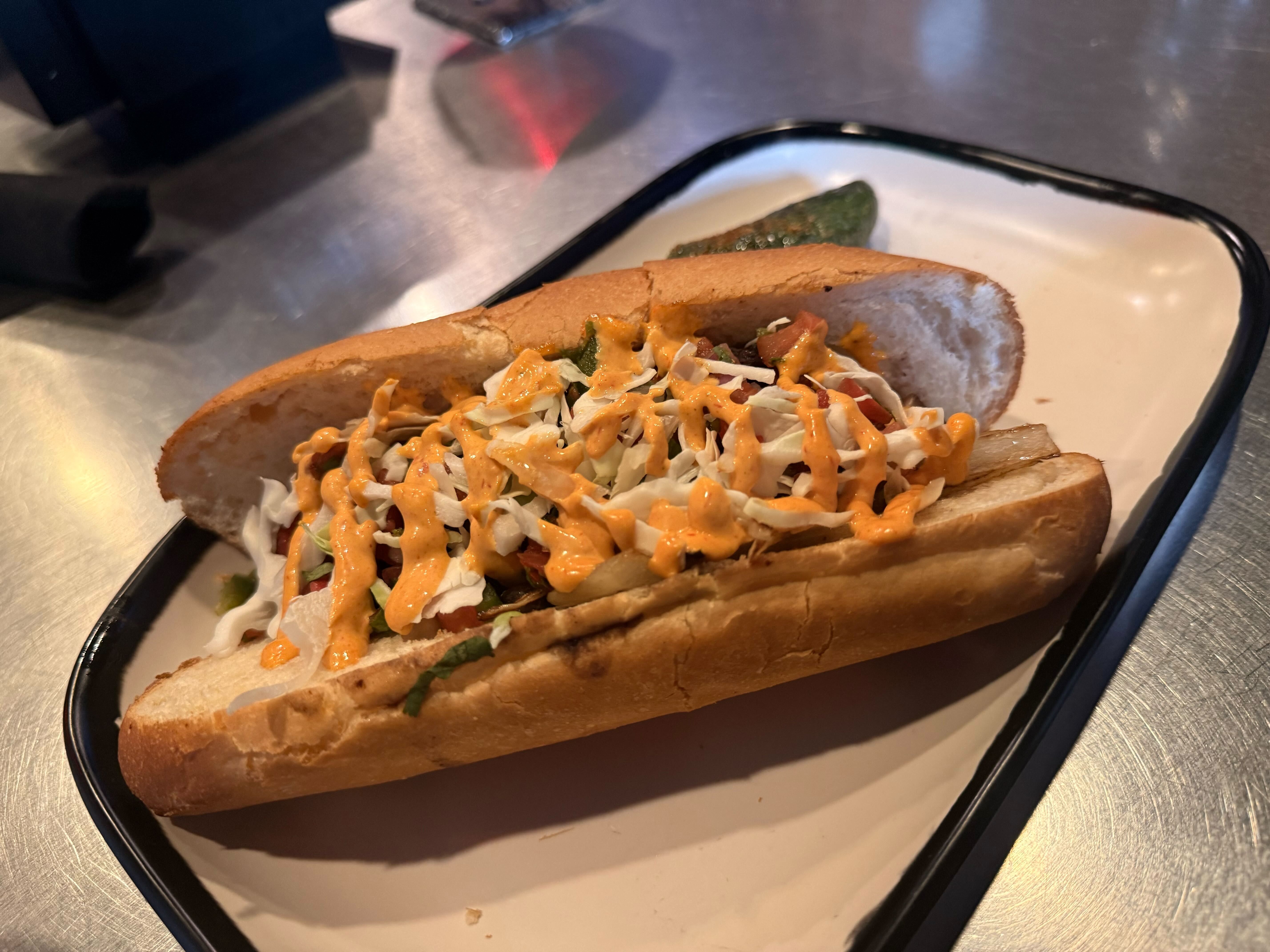 Hot dog in a crusty bun topped with shredded cabbage slaw and orange mayo drizzle; served on a white enamel plate with a pickle spear nearby.