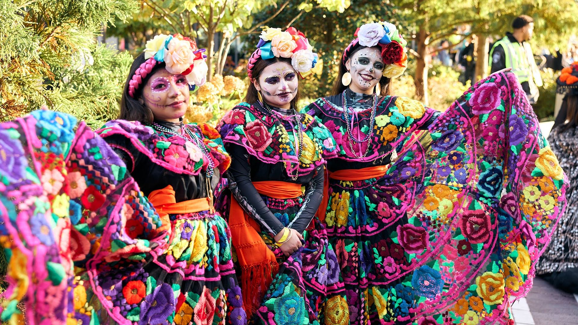 Three women in bright, embroidered dresses with large flower headpieces and sugar skull makeup pose outdoors at a sunny festival, surrounded by green trees.
