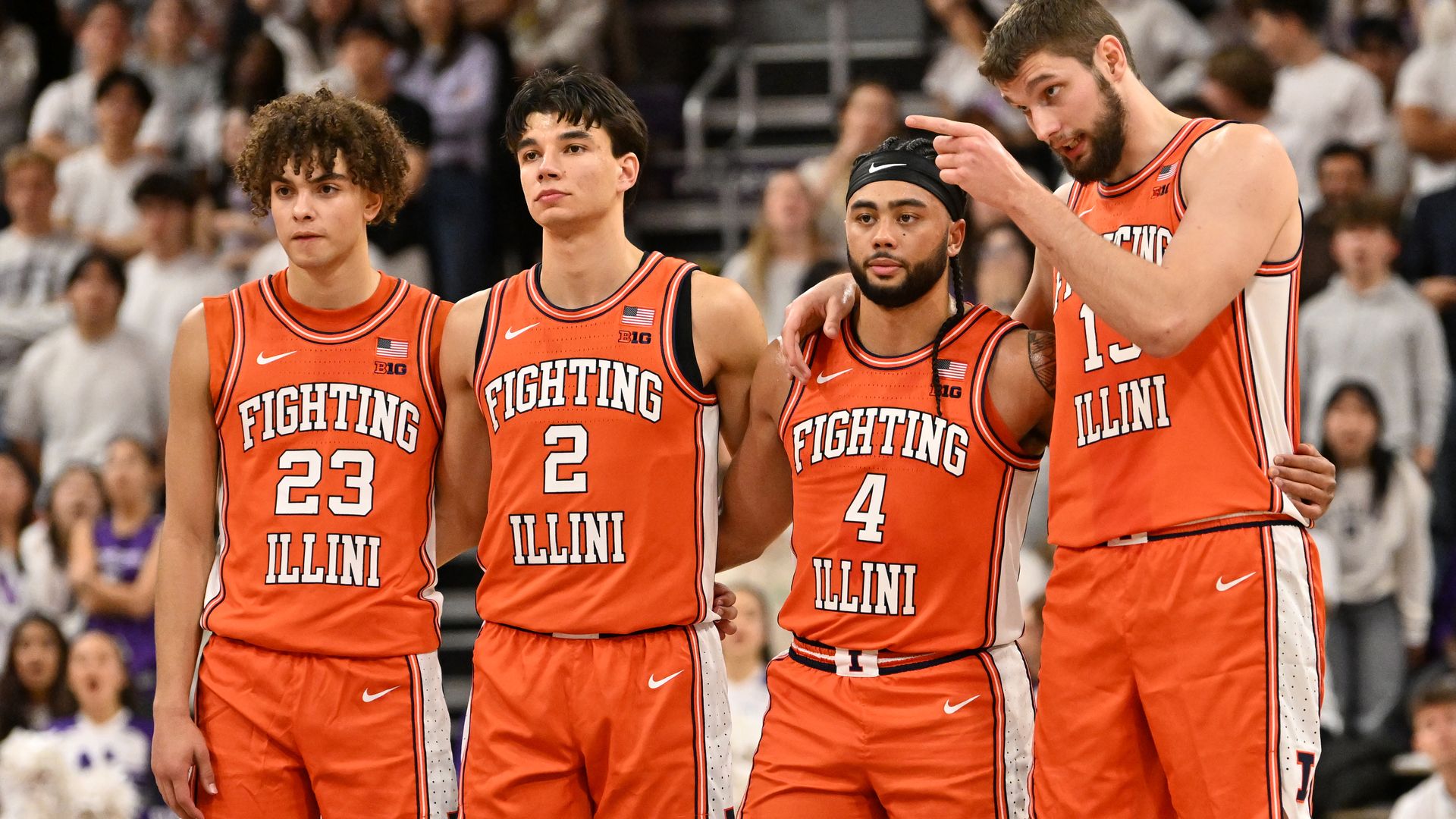 Four Illinois Fighting Illini basketball players stand together on the court in orange uniforms with white trim, 'FIGHTING ILLINI' on their jerseys, arms around each other as a crowd watches.