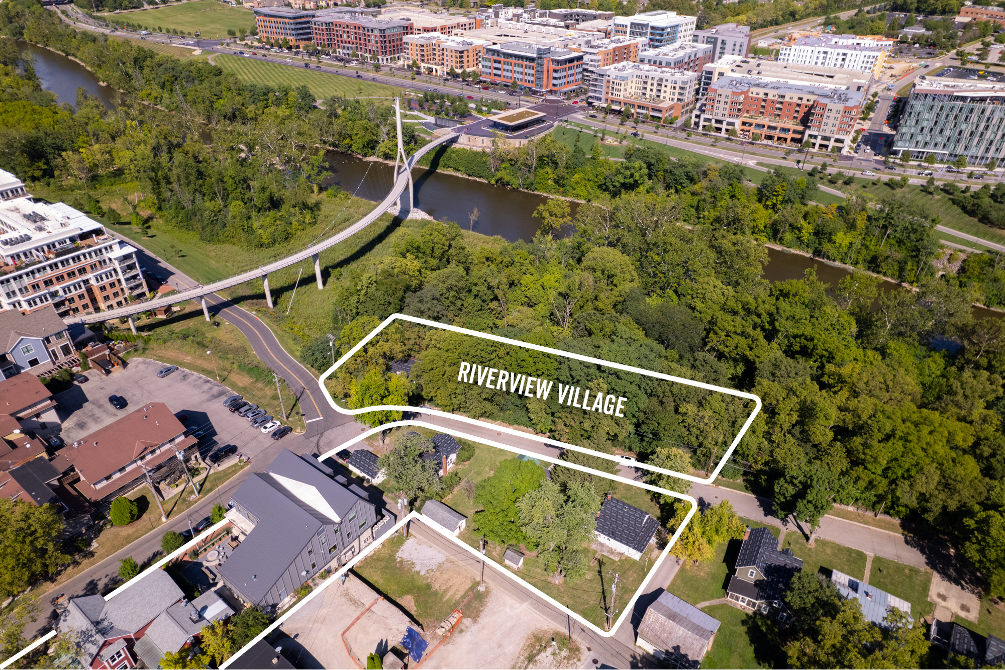 Aerial view of Riverview Village outlined in white, surrounded by dense green trees, a river with a pedestrian bridge, roads, and mixed residential and commercial buildings.