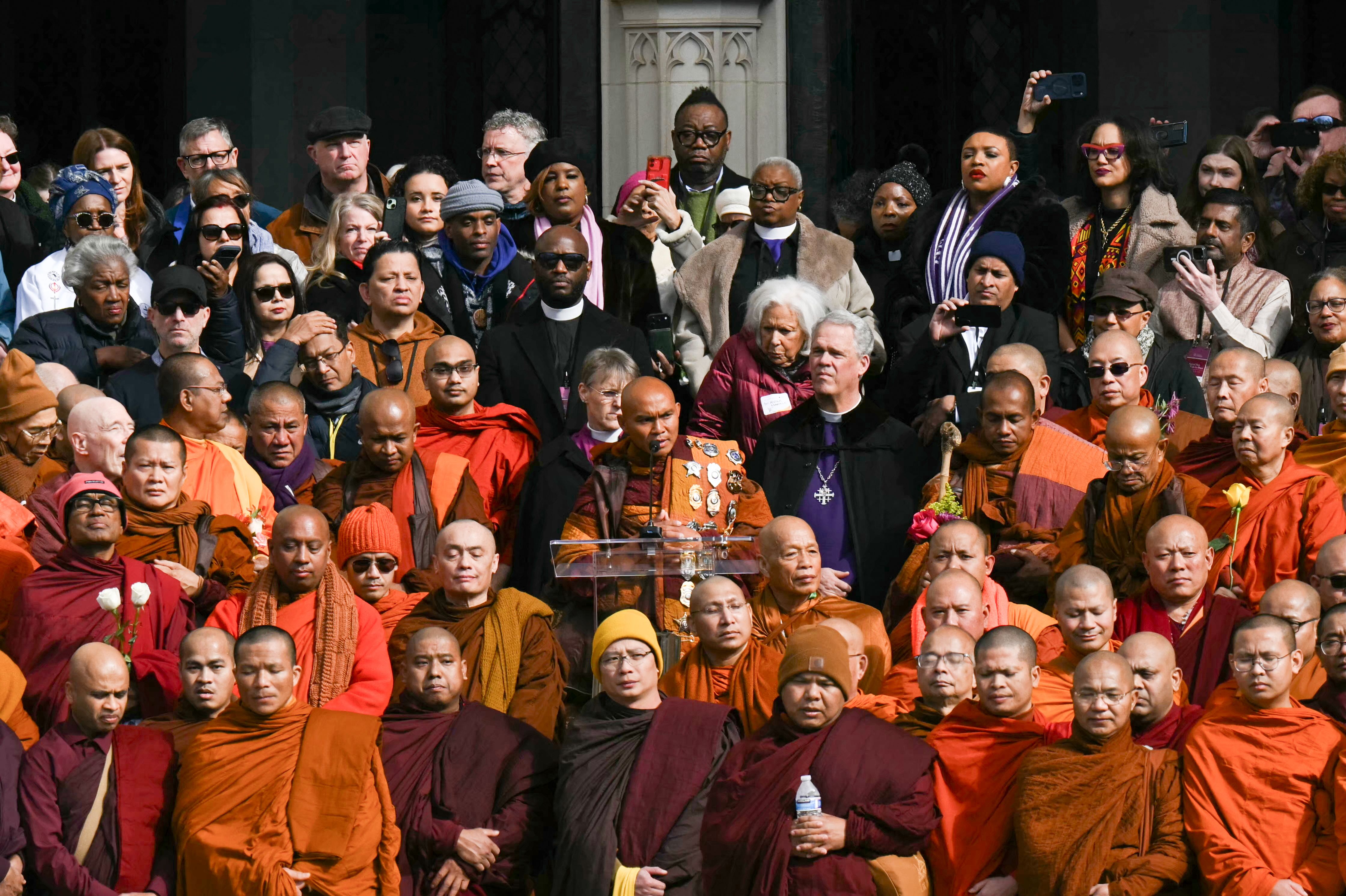 Large diverse group including Buddhist monks in orange and maroon robes and others gathering outdoors, some using phones to take pictures, monk speaking at clear podium.