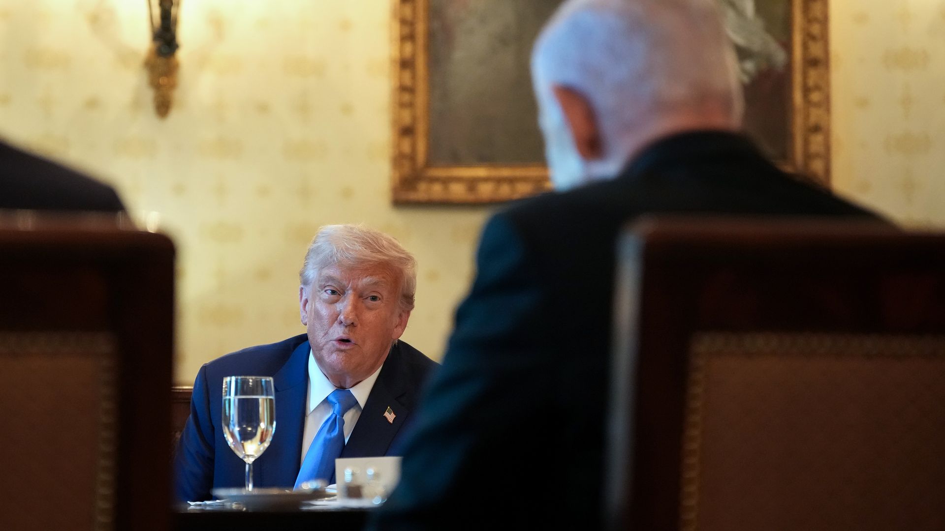 Man in a blue suit and blue tie speaking at a formal dining table with glass of water and dishes, another person with white hair partially visible in foreground, ornate wall decor in background.