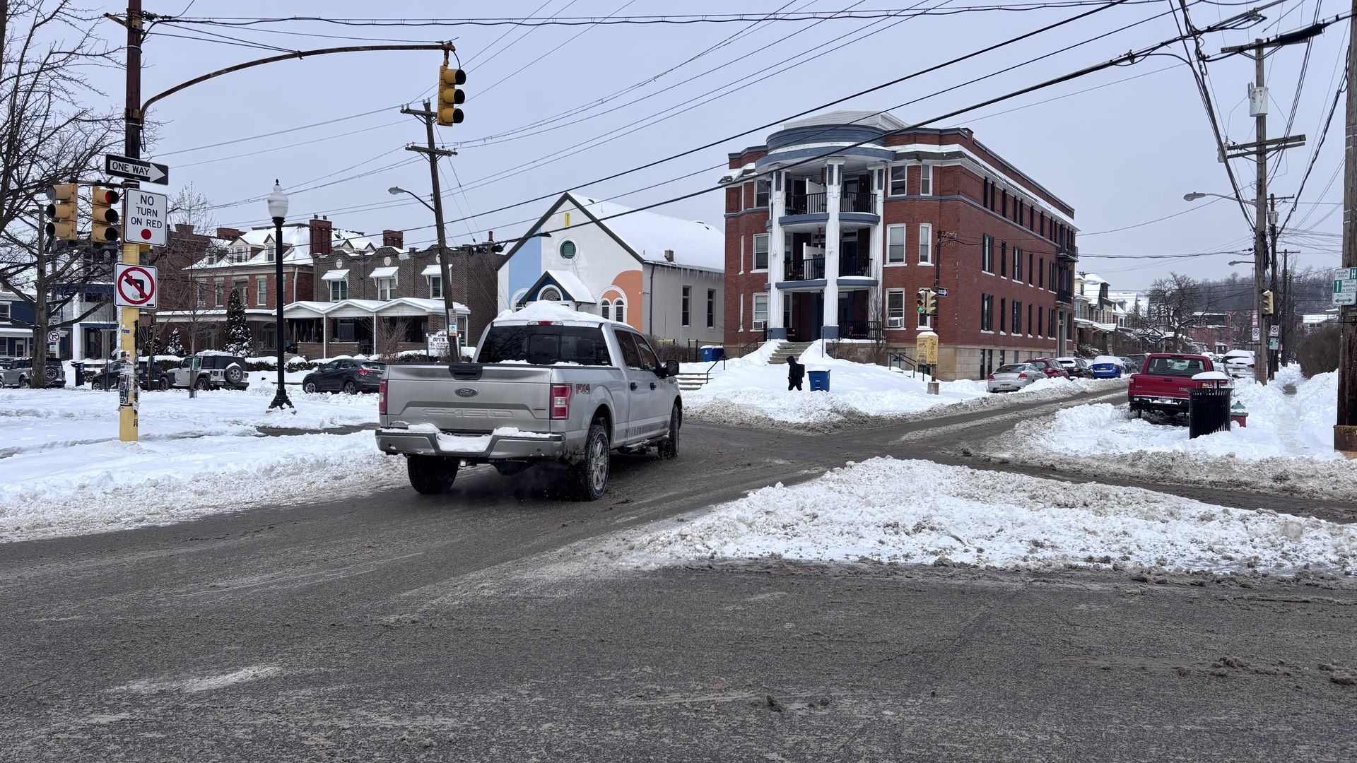 Snow-covered street intersection with cars, traffic lights, and a large brick building with white columns on the corner under a gray sky.
