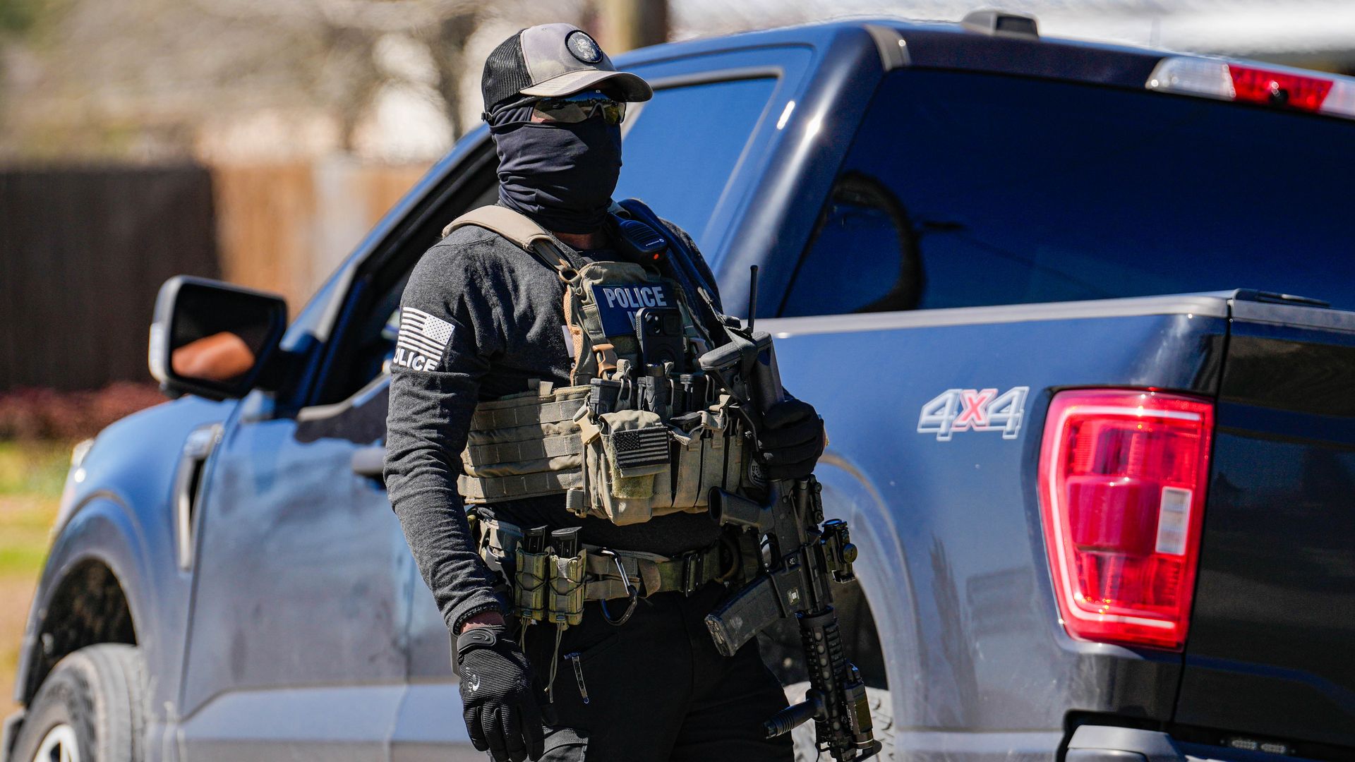 Police officer in tactical gear, black mask, and gloves standing next to a dark blue 4x4 pickup truck, holding a rifle. Daylight outdoor setting with blurred background.