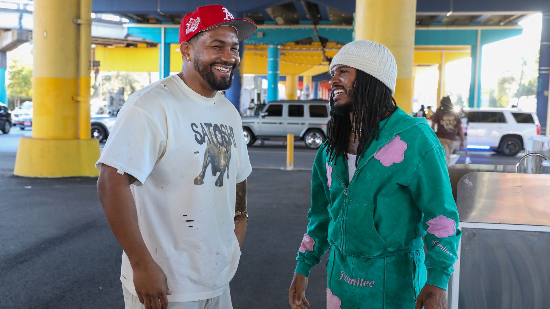 Two men smiling and talking under a colorful overpass; one wears a white shirt with a red cap, the other wears a green tracksuit with pink clouds and a white beanie.