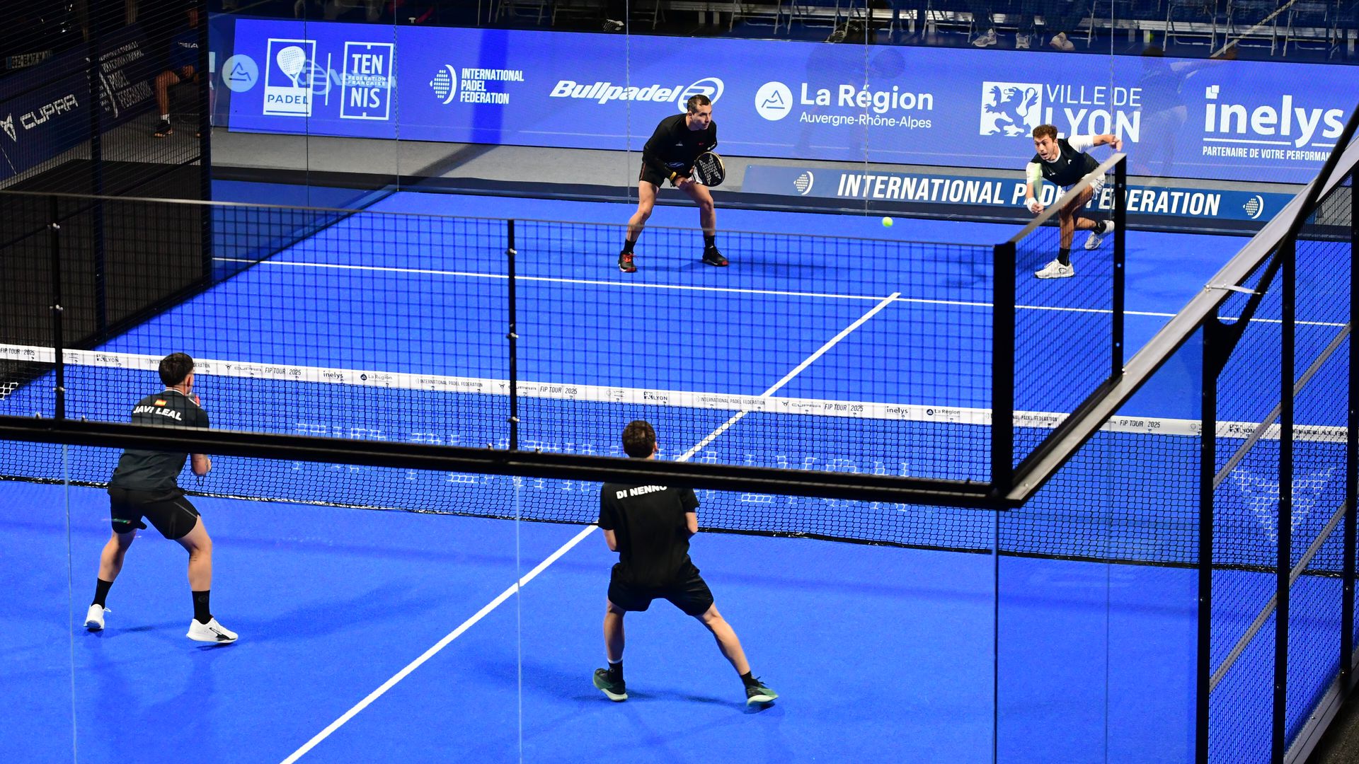 Four men in black exercise clothing stand on a blue, tennis-style court, with two on each side of the net; the court is surrounded by glass walls. 
