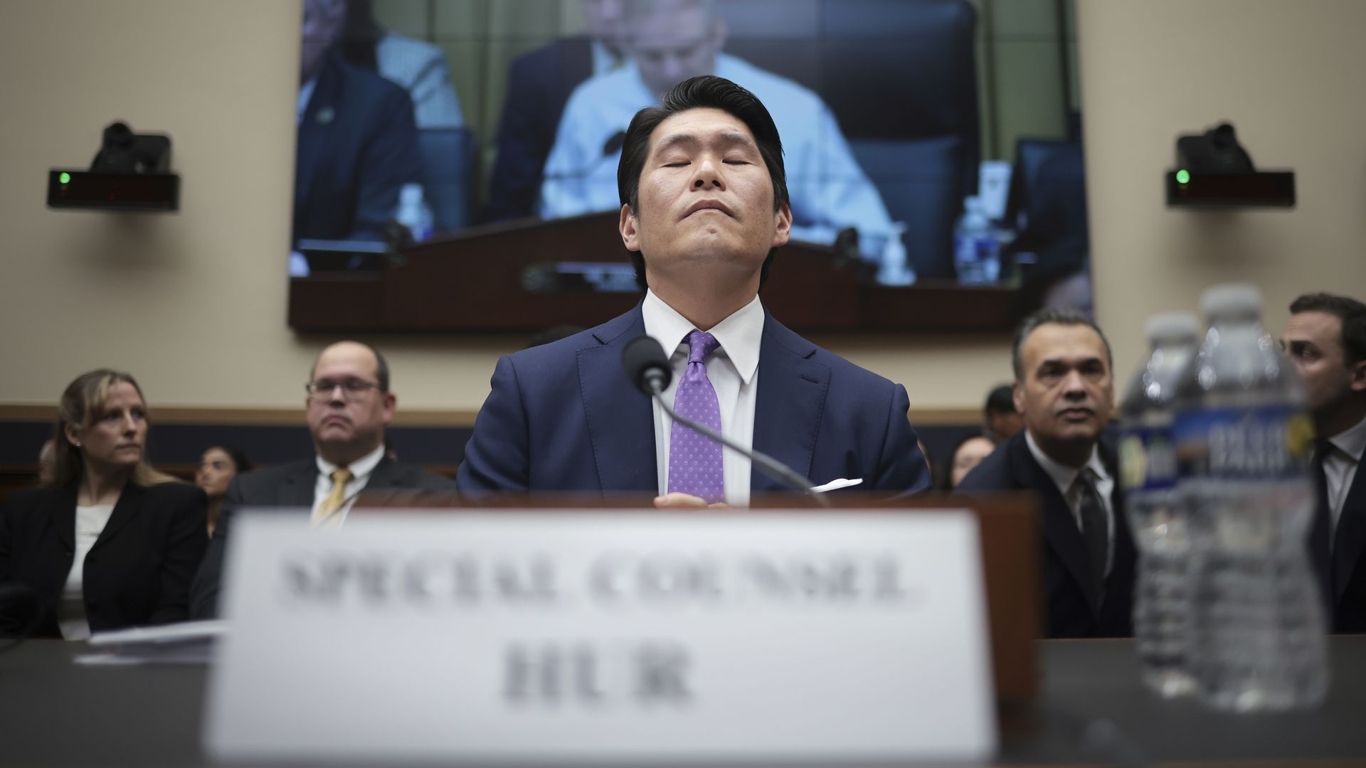 Former Biden special counsel Robert Hur, wearing a dark blue suit, white shirt and purple tie, sits at a microphone in front of a large flat screen TV with Rep. Jim Jordan on it.