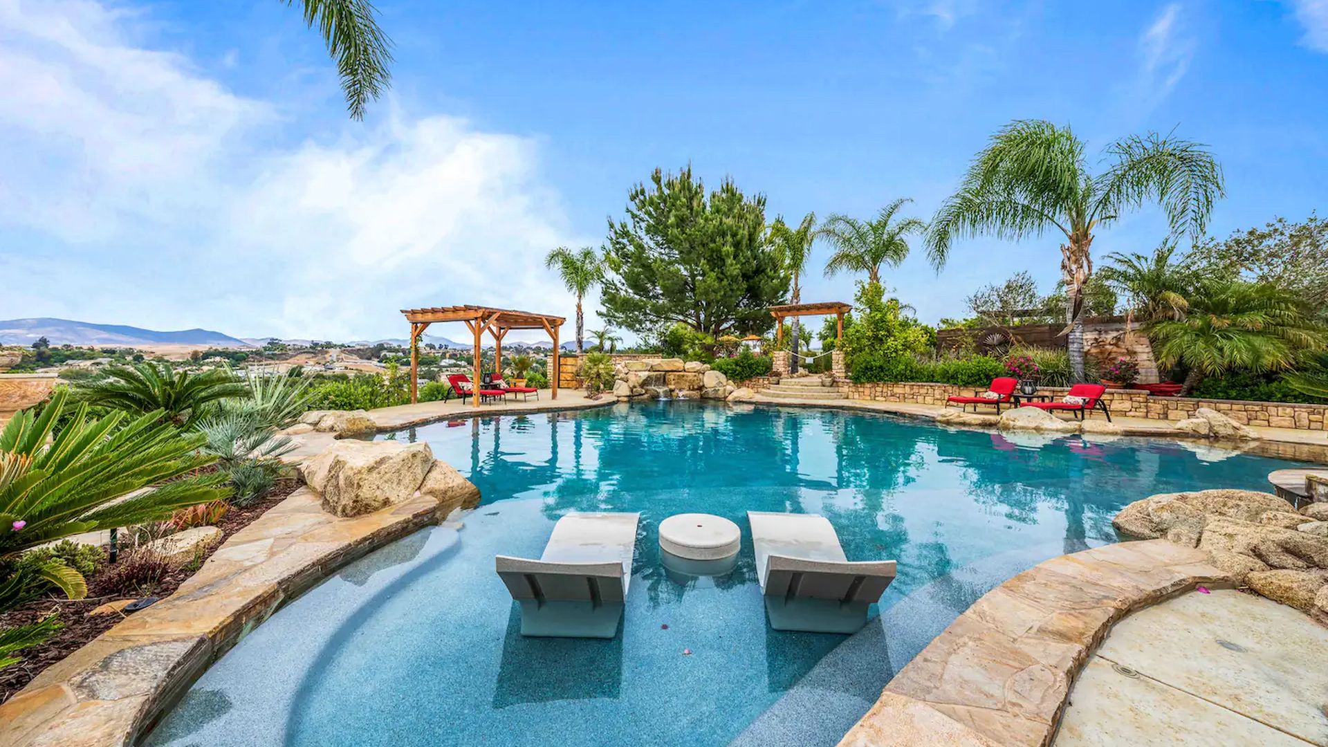 The view from two lounge chairs in a resort-style pool features palm trees,  vineyards and desert mountains. 