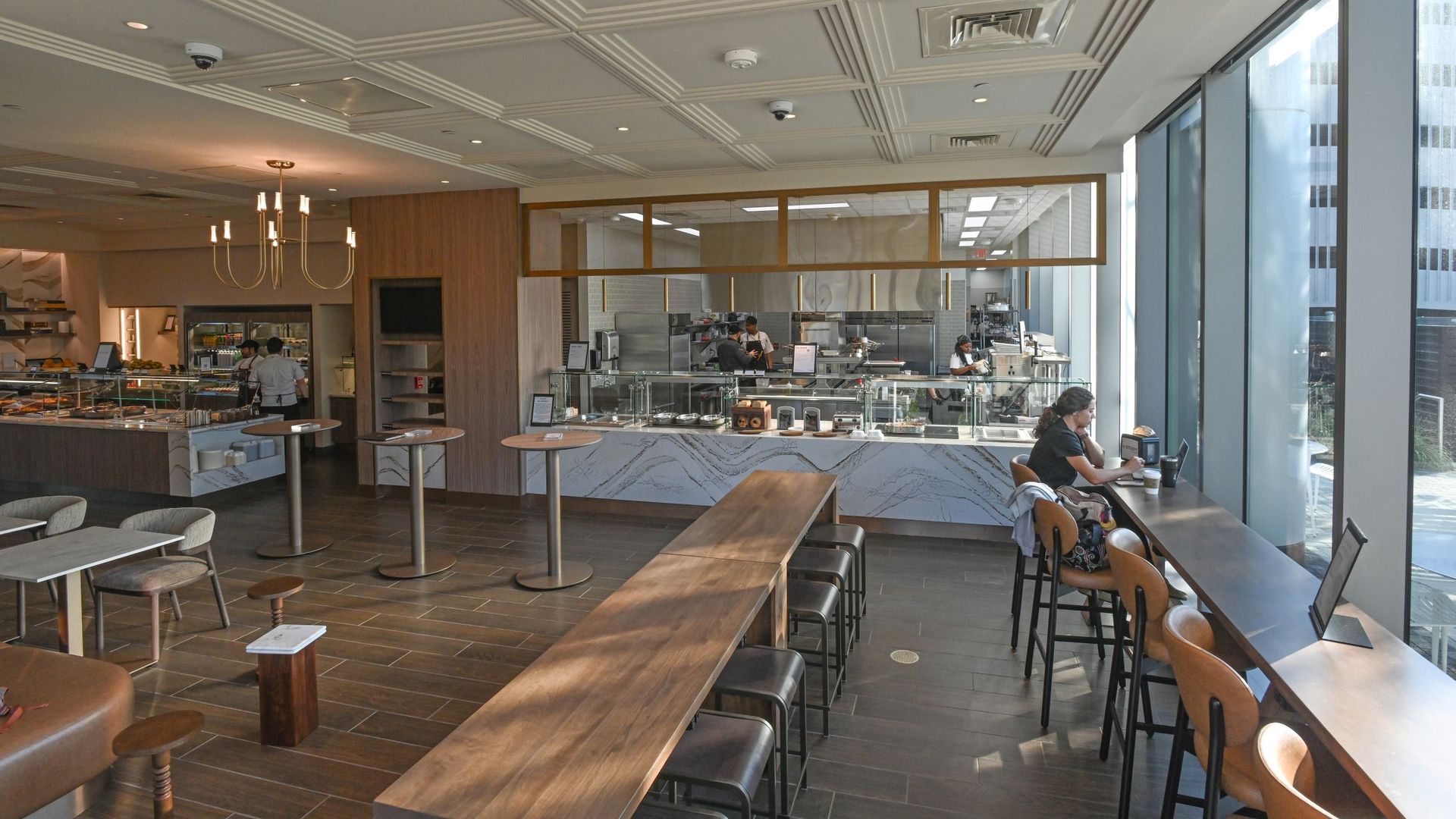 Bright cafe interior with long wooden tables and stools, a glass pastry counter, staff behind it, and large windows along the right; paneled ceiling and warm chandeliers.