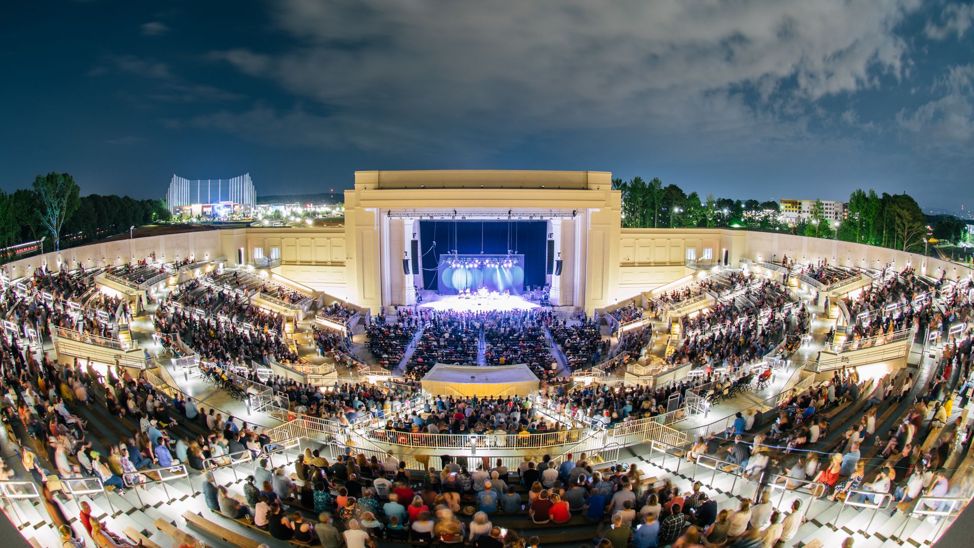 Night view of a large outdoor amphitheater with tiered seating filled with spectators watching a brightly lit stage performance under a partly cloudy sky.