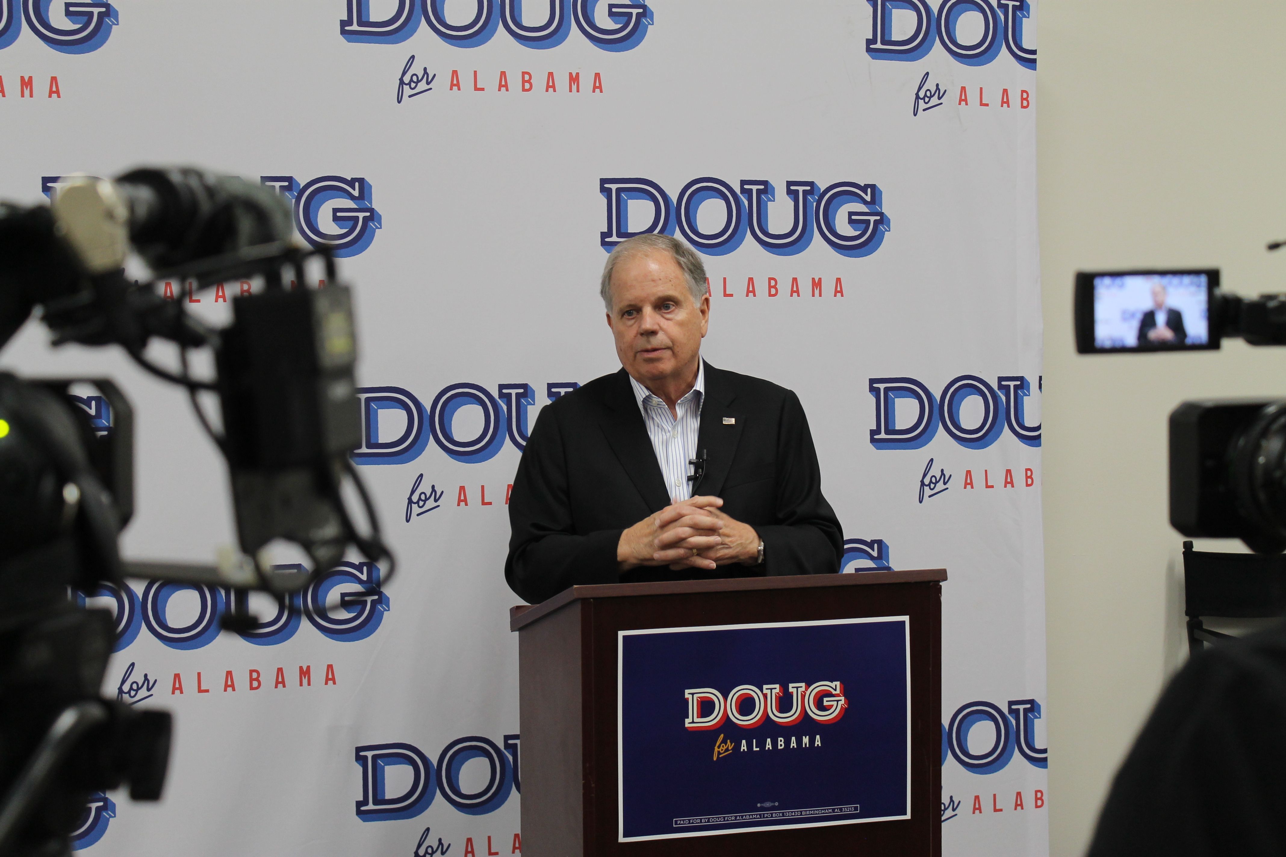 A man in a dark suit stands at a podium for a press conference, surrounded by cameras; the backdrop repeats blue DOUG logos with red ALABAMA text.
