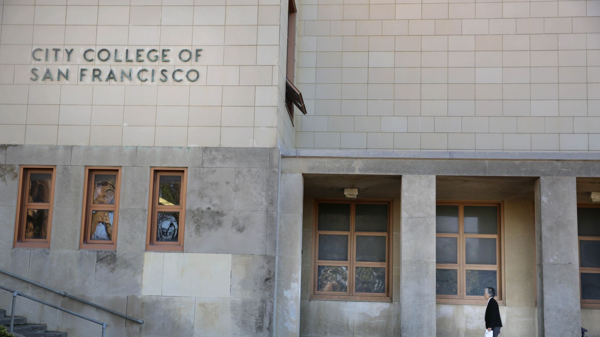 Exterior of City College of San Francisco with beige tiled walls, small windows, stairs on left, and a person pulling a suitcase walking under a sheltered area.