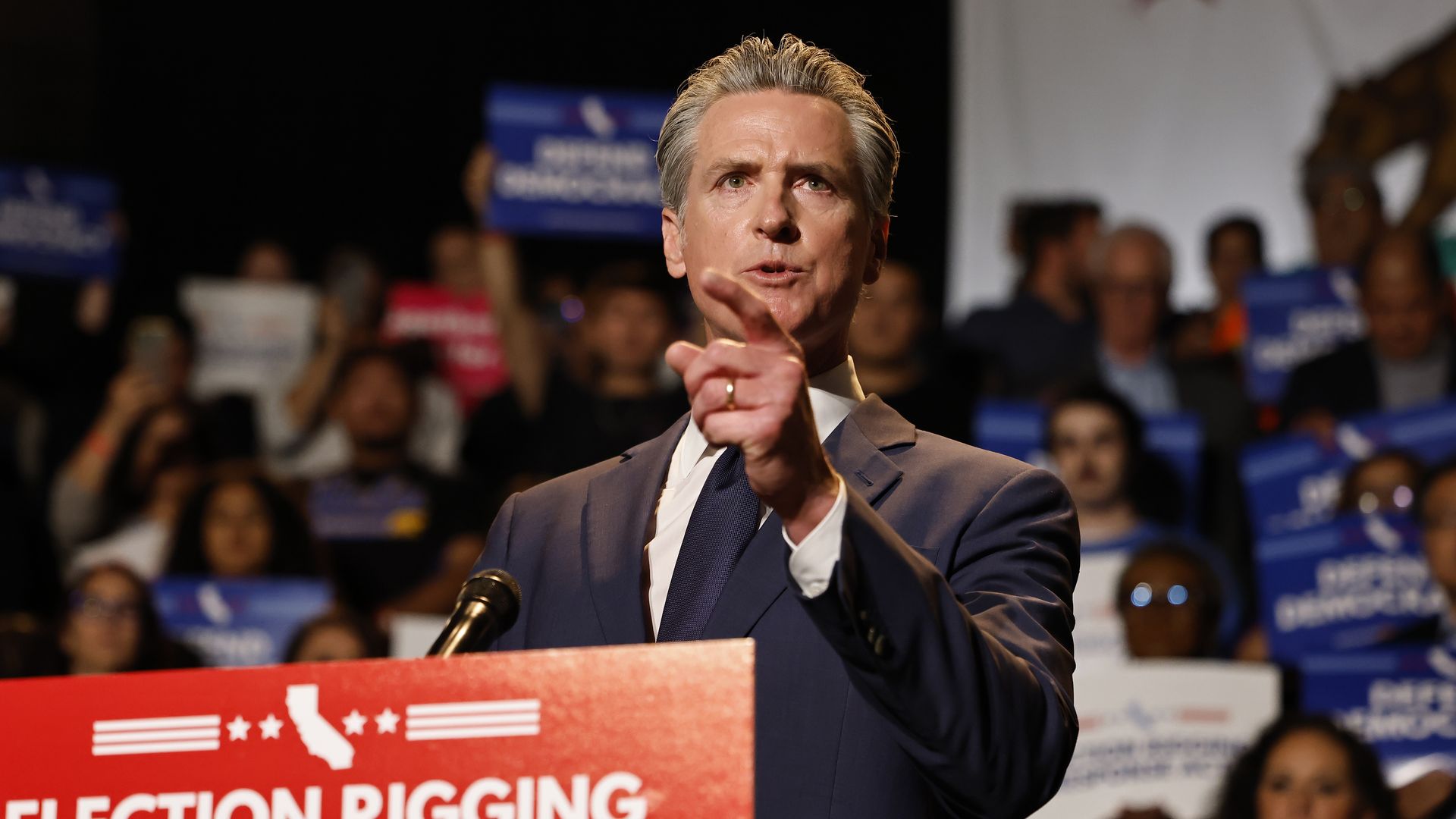 Man in a dark suit speaking passionately at a podium with a red sign reading "ELECTION RIGGING RESPONSE ACT," pointing forward, with a crowd holding blue signs blurred in the background.