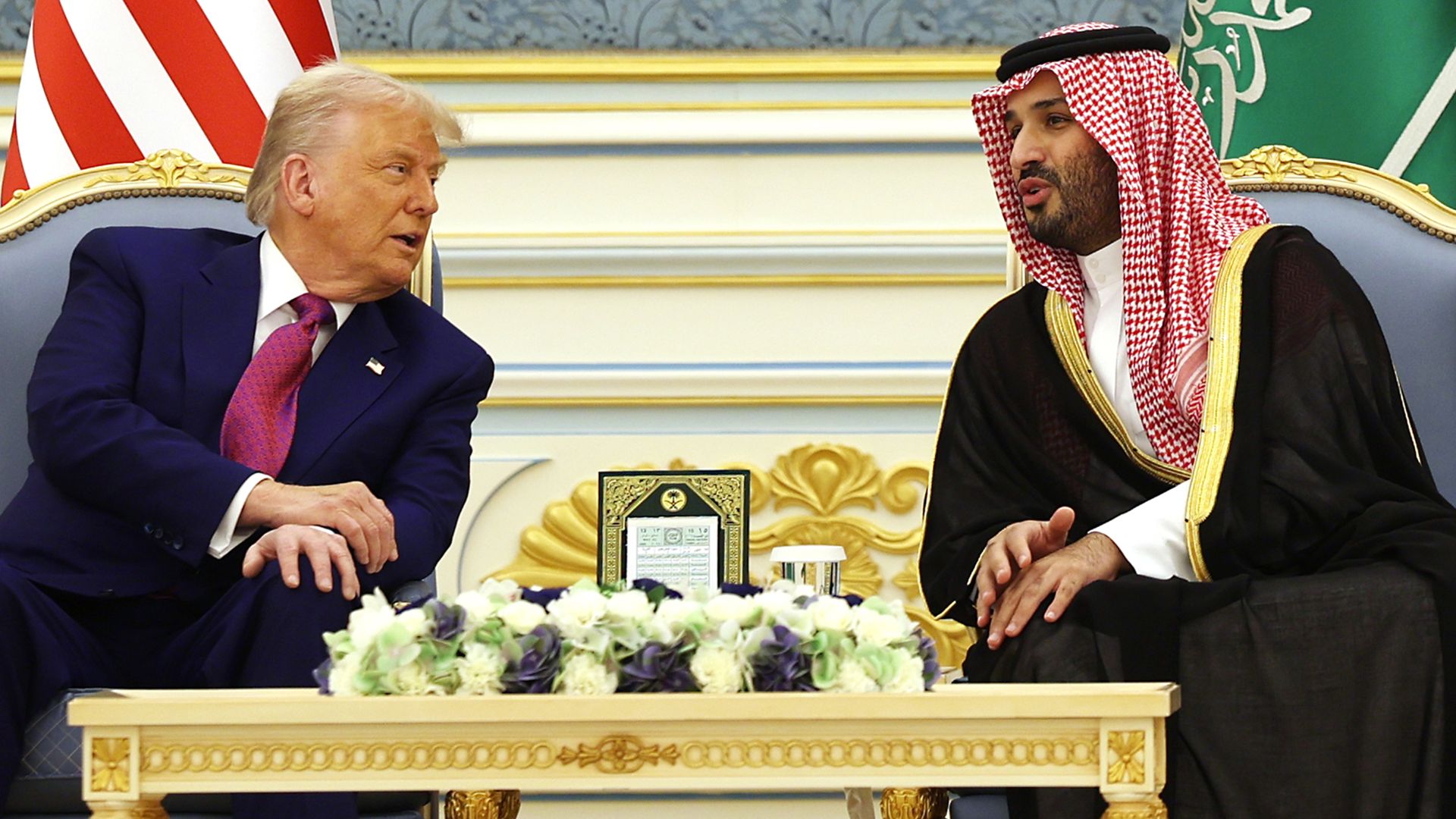 Two men in a formal meeting sit on gold-trimmed chairs across a table; one in a blue suit with pink tie, the other in traditional Arab attire with a red-and-white keffiyeh; US and Saudi flags behind.