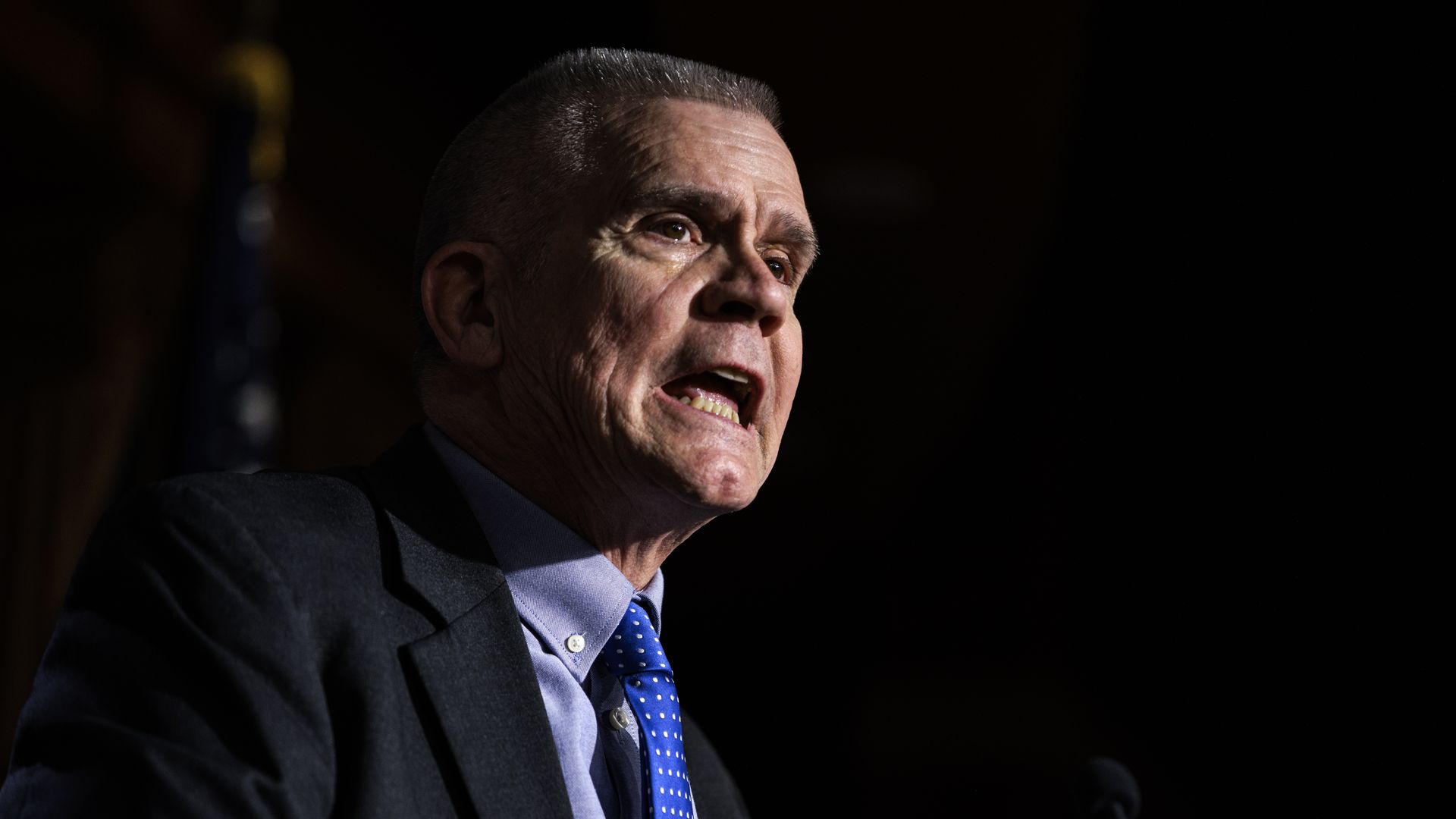 Rep. Matt Rosendale, wearing a blue suit and standing in front of an American flag against a dark backdrop.