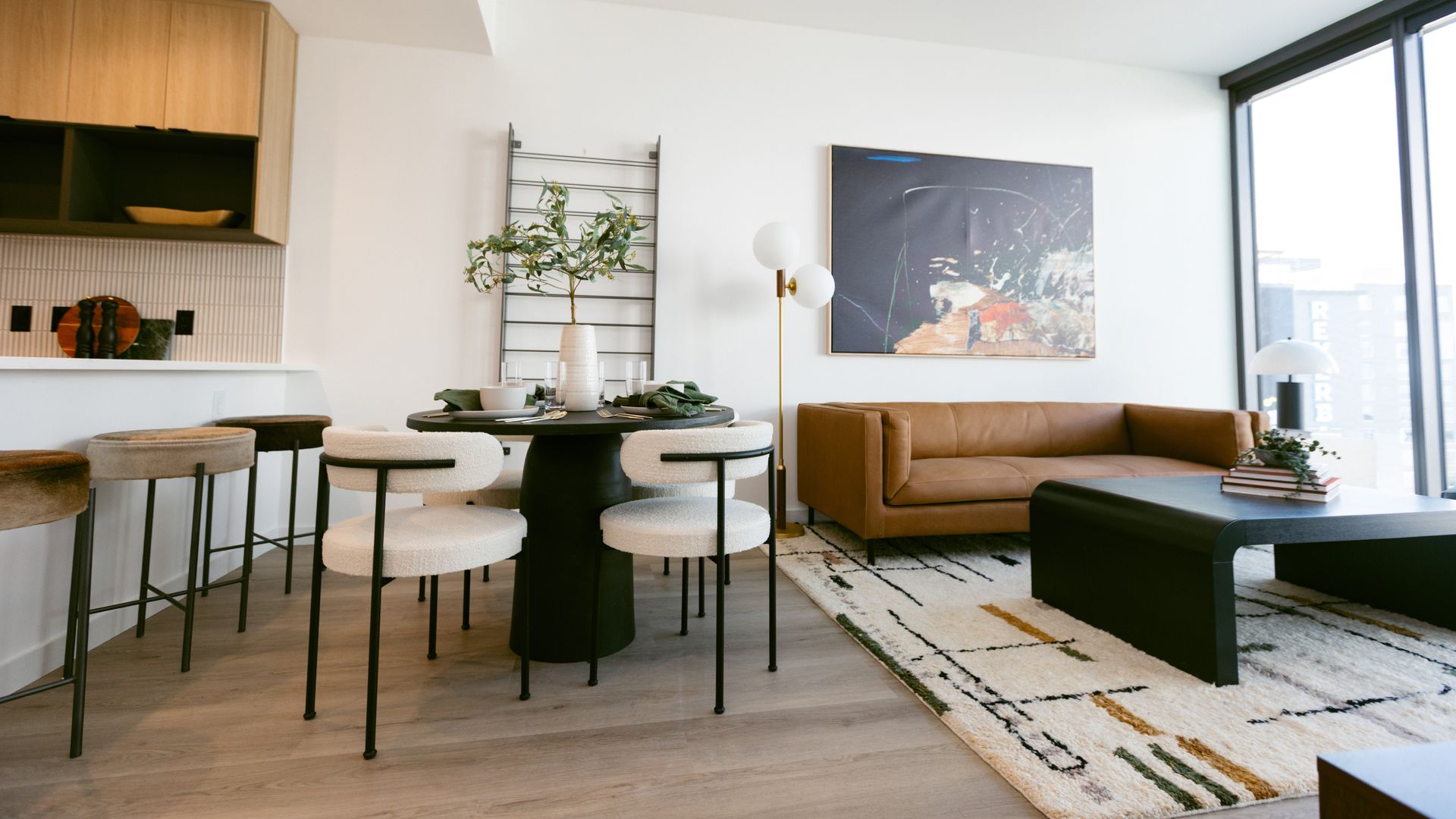 A modern living room and dining area with tan leather sofa, black round dining table with white chairs, abstract art, floor lamp, large window, and light wood flooring.