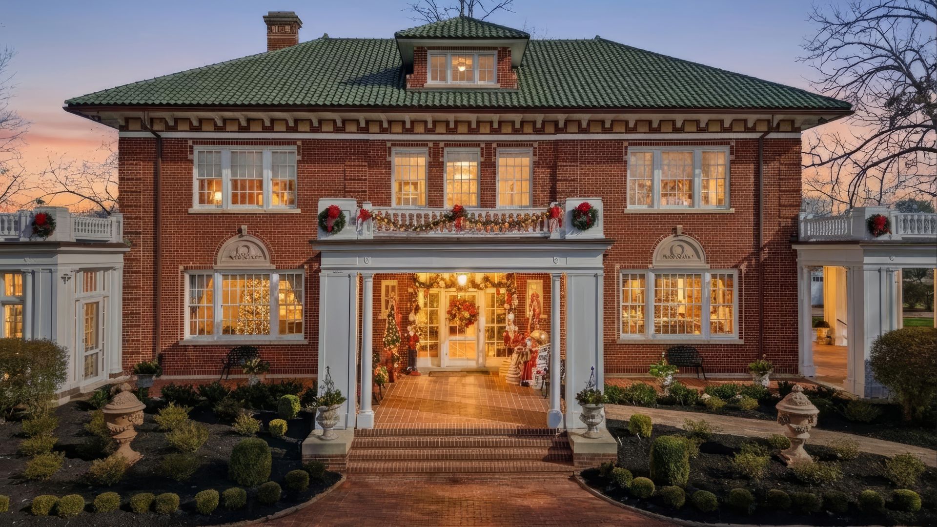 Twilight view of a large red brick house with green roof and white pillars, decorated with Christmas wreaths and garlands, warm lights glowing inside, and manicured shrubs in front.