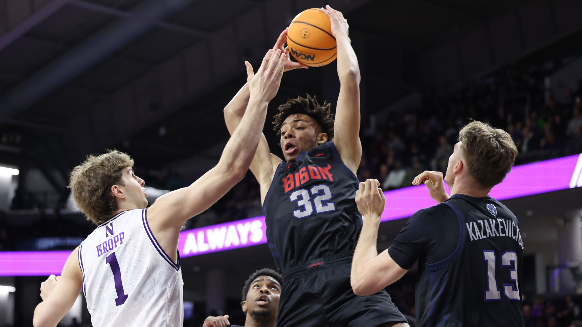 Basketball action: a dark Bison jersey player (32) leaps to shoot, contested by a white-jersey defender (1) and a dark-jersey defender (13); orange ball, crowd and purple arena signage in the background.